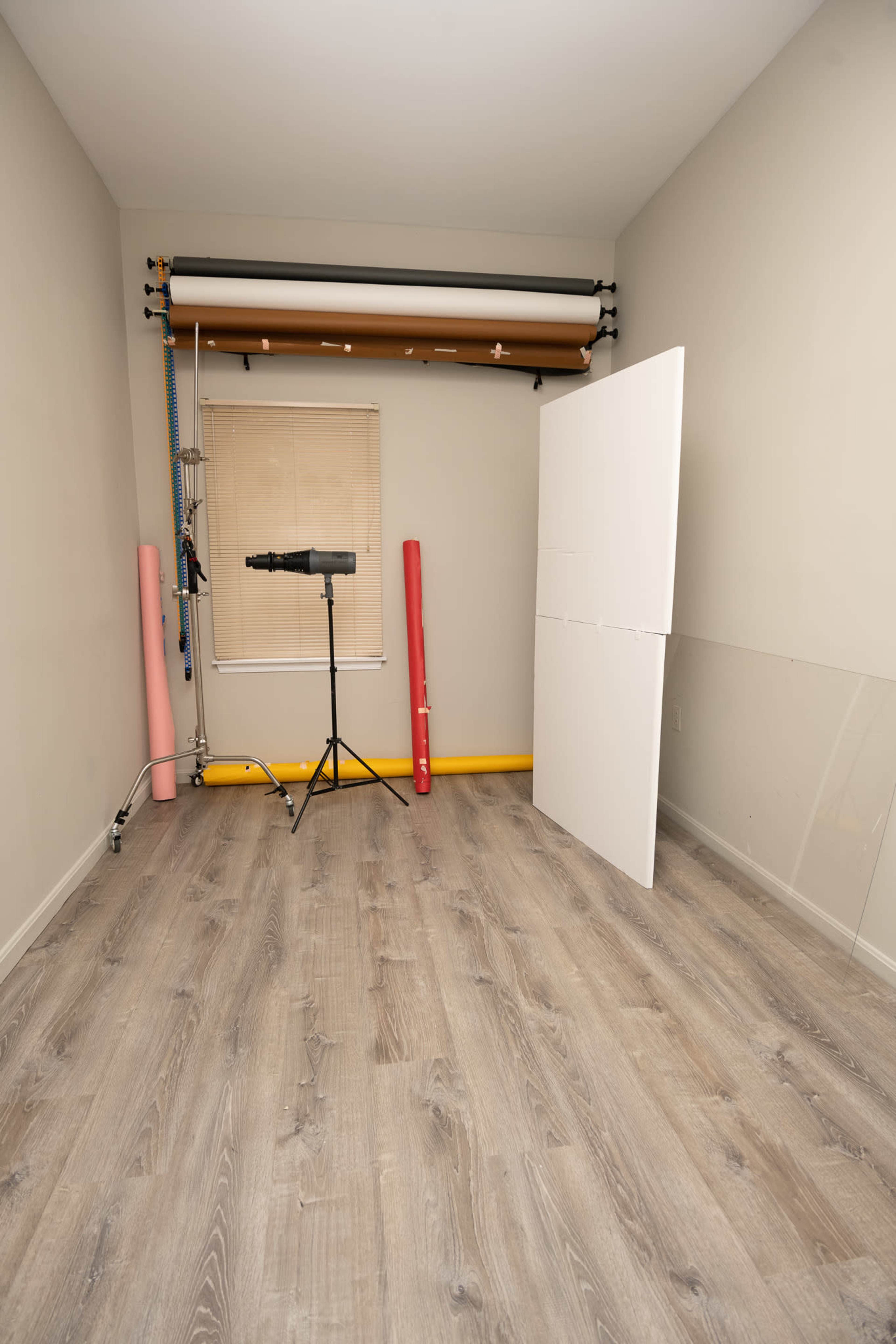 The image shows a photography studio with a backdrop setup, a light stand, and rolled paper in pink and yellow against a gray wall and floor.