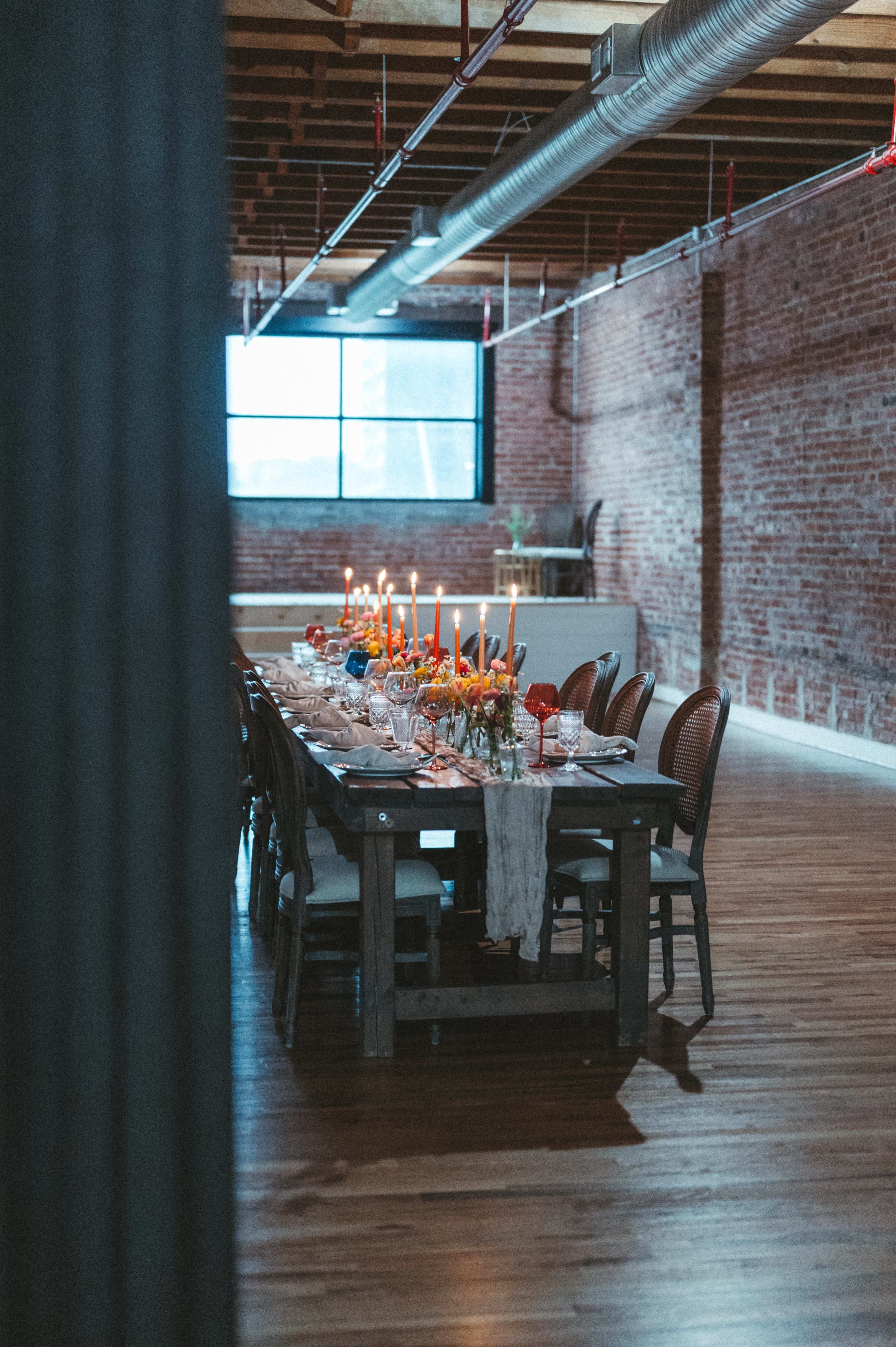A long dining table with candles and flower centerpieces is set in a room with exposed brick walls and large windows.