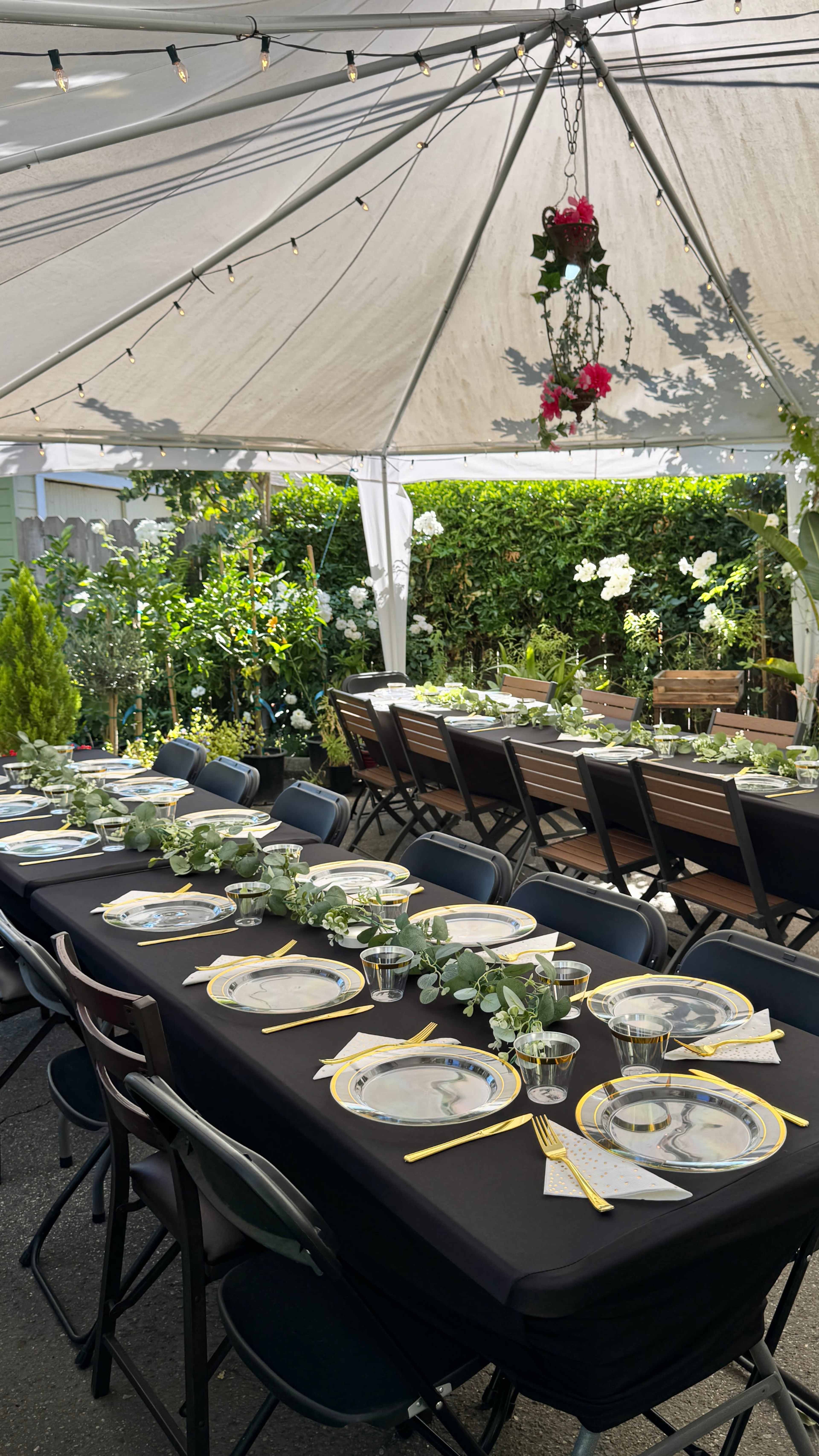 A large outdoor dining setup features long tables decorated with black tablecloths, plates, and greenery under a tent with string lights.
