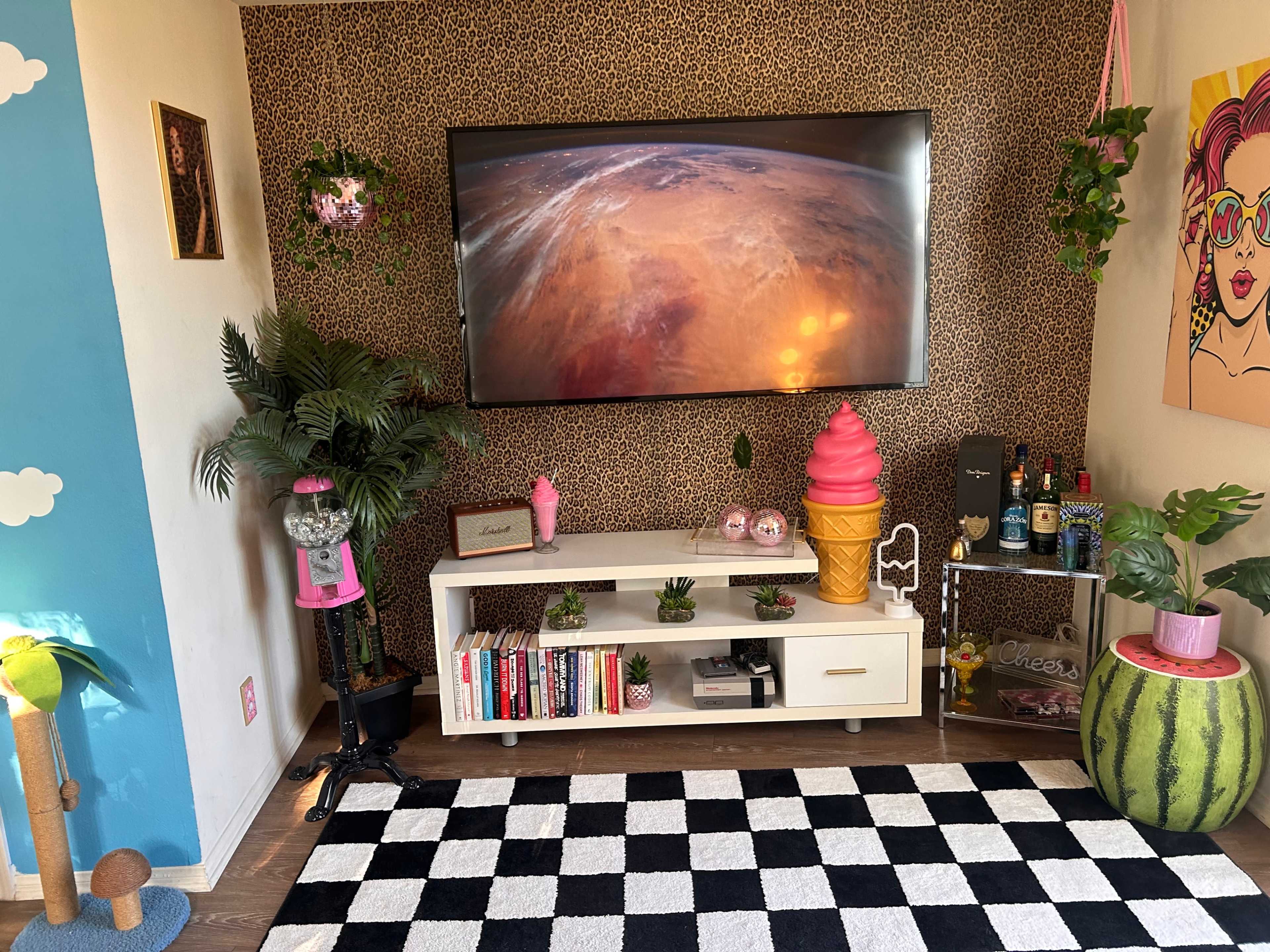 The room features a large TV mounted on a leopard-print accent wall, with a white entertainment unit displaying books and decorative items, surrounded by potted plants and colorful décor.