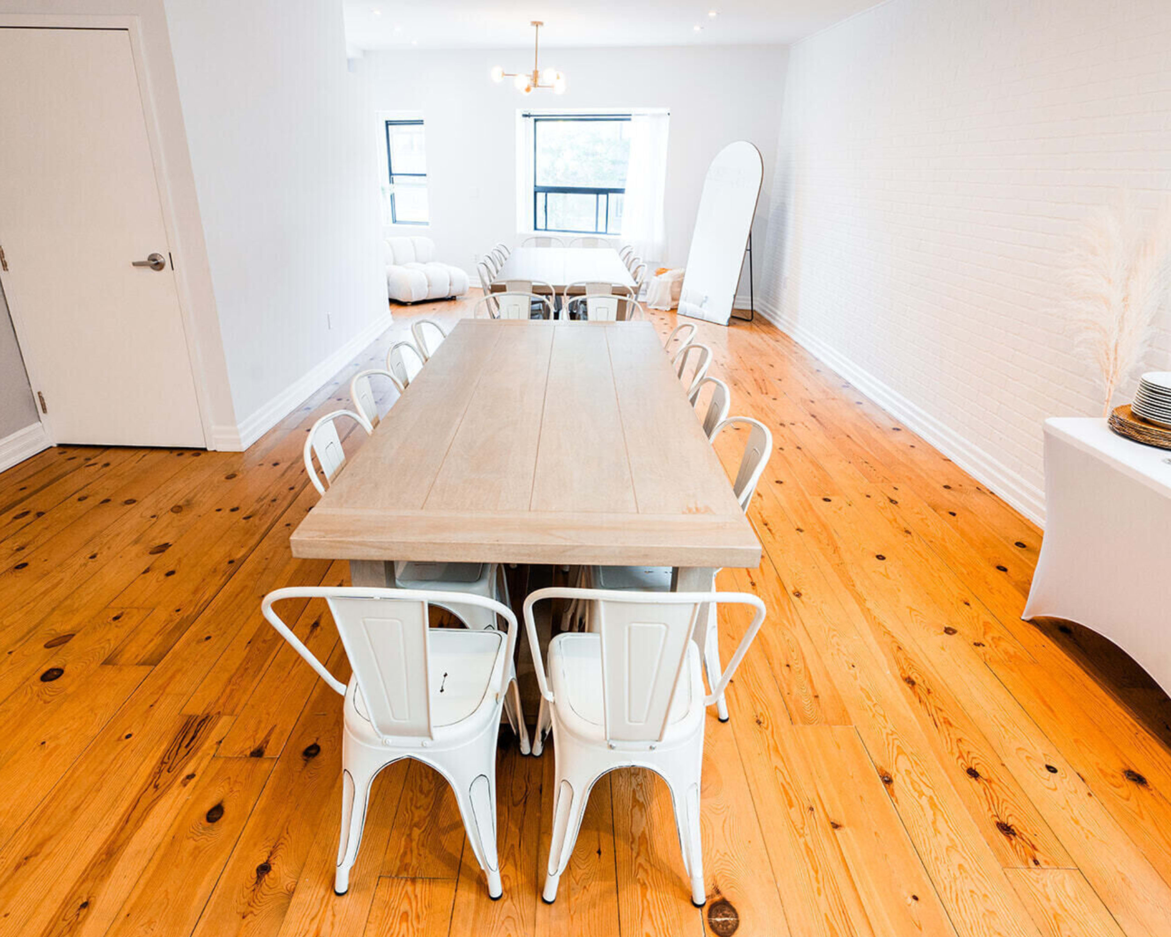 A large wooden dining table surrounded by white metal chairs in a well-lit, minimalistic room with hardwood flooring.
