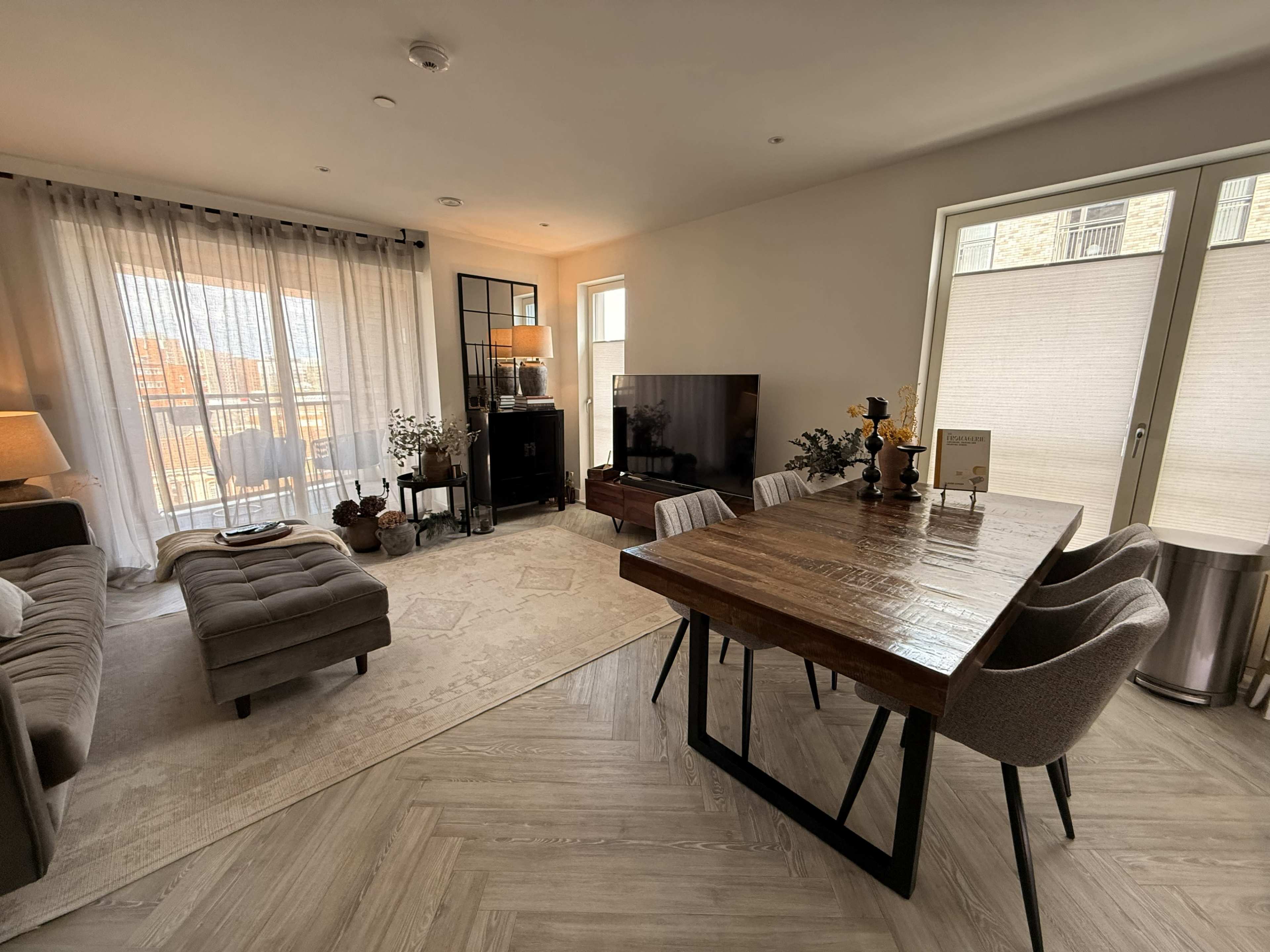 The living room features a wooden dining table surrounded by chairs, a large television, a couch, and large windows covered with sheer curtains.
