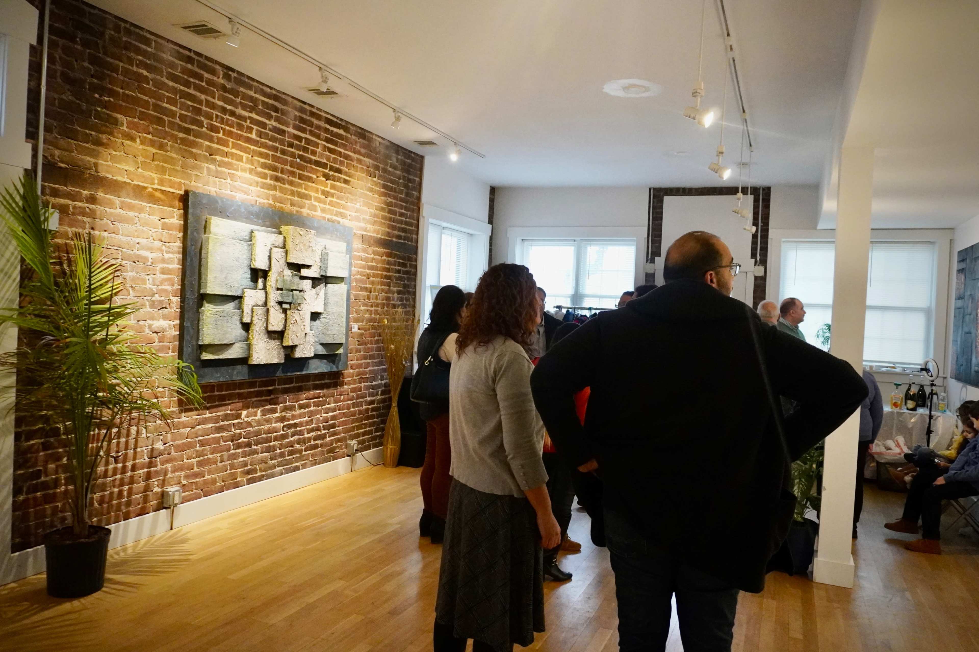 The scene shows a group of people gathered in a gallery space with a textured artwork on a brick wall.