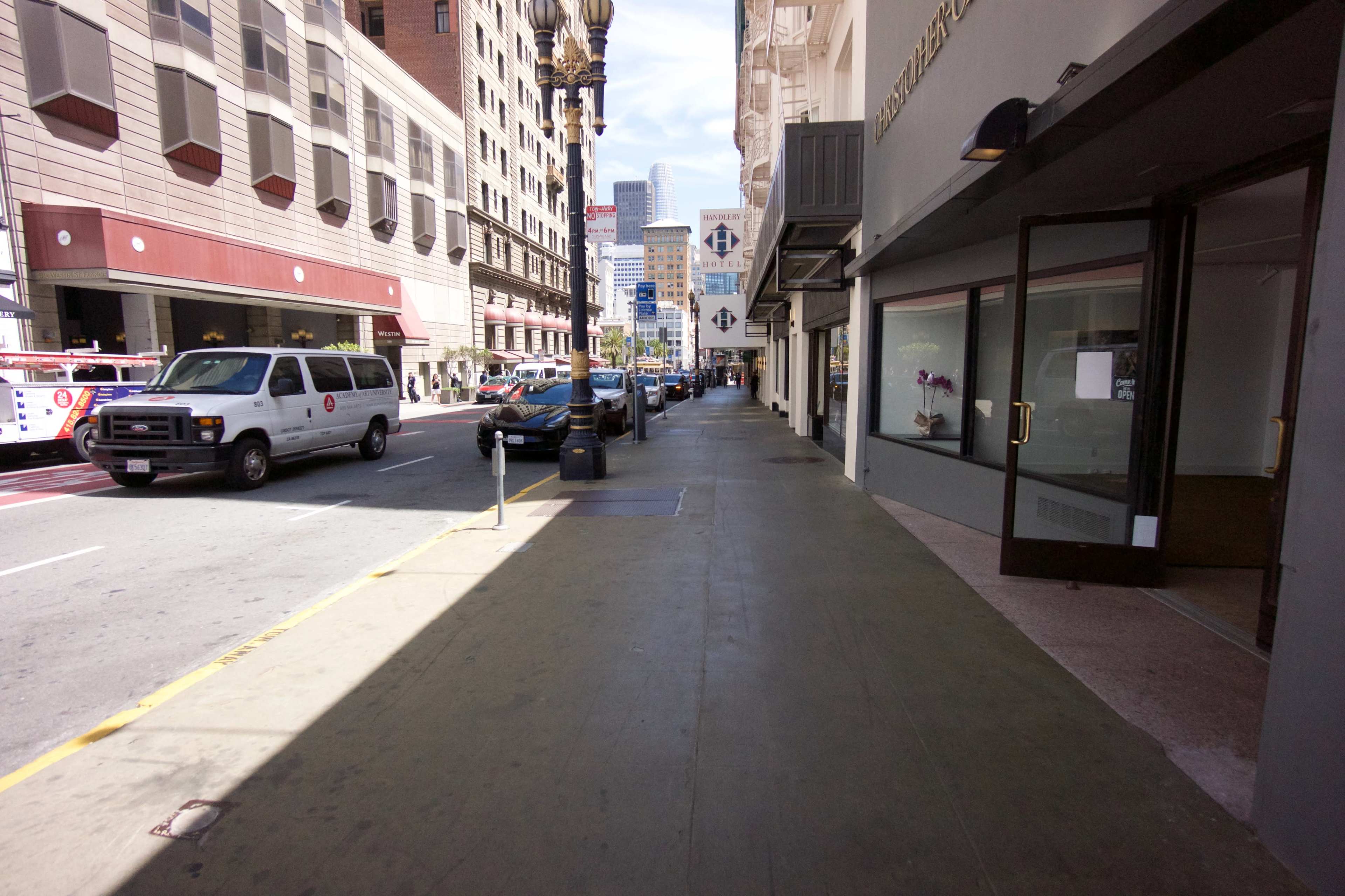 The image shows a wide sidewalk adjacent to a street with parked cars and several buildings lining the road.