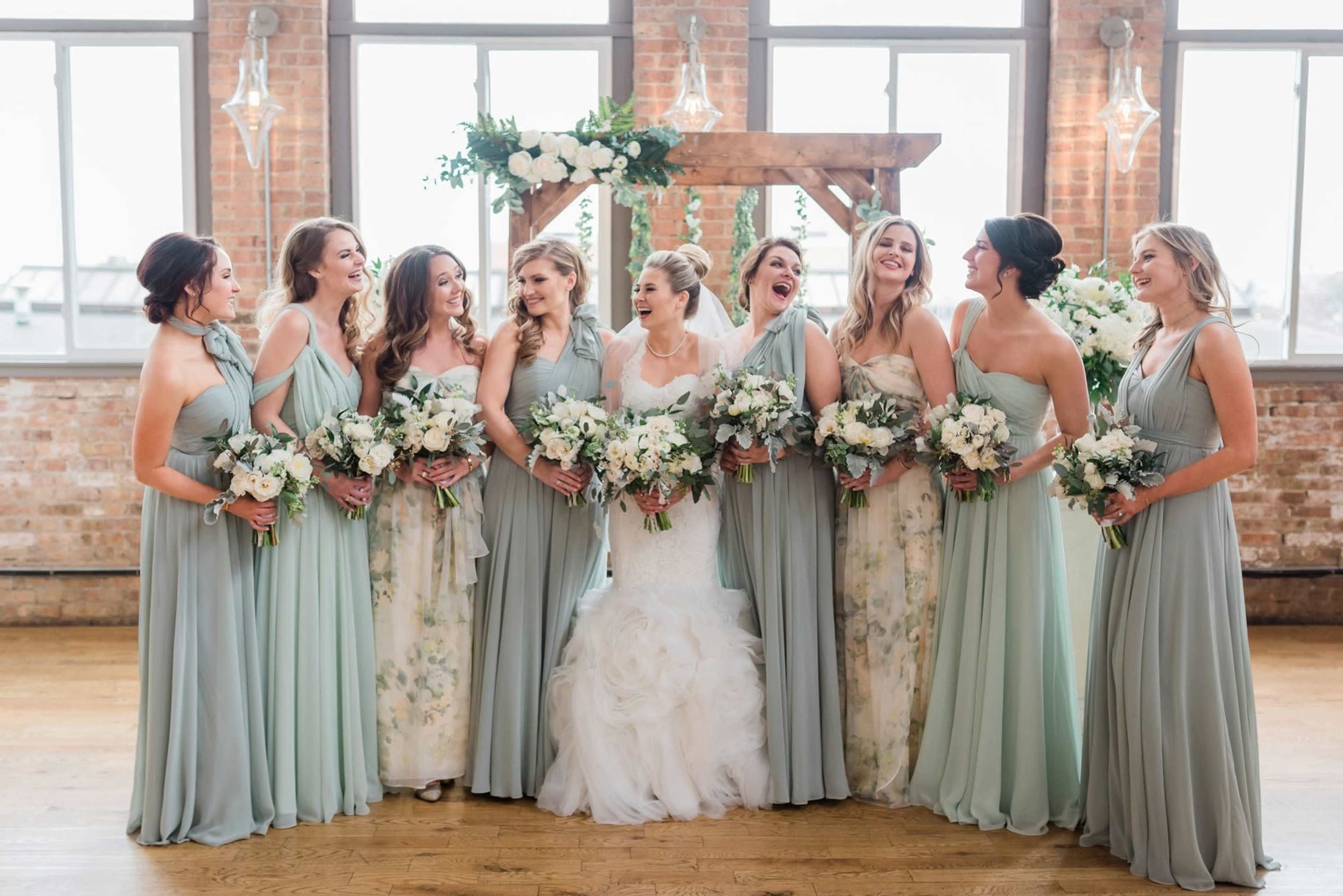 A bride wearing a white gown stands in the center of a group of nine bridesmaids in pastel dresses, all smiling and holding flower bouquets in a well-lit, rustic venue.
