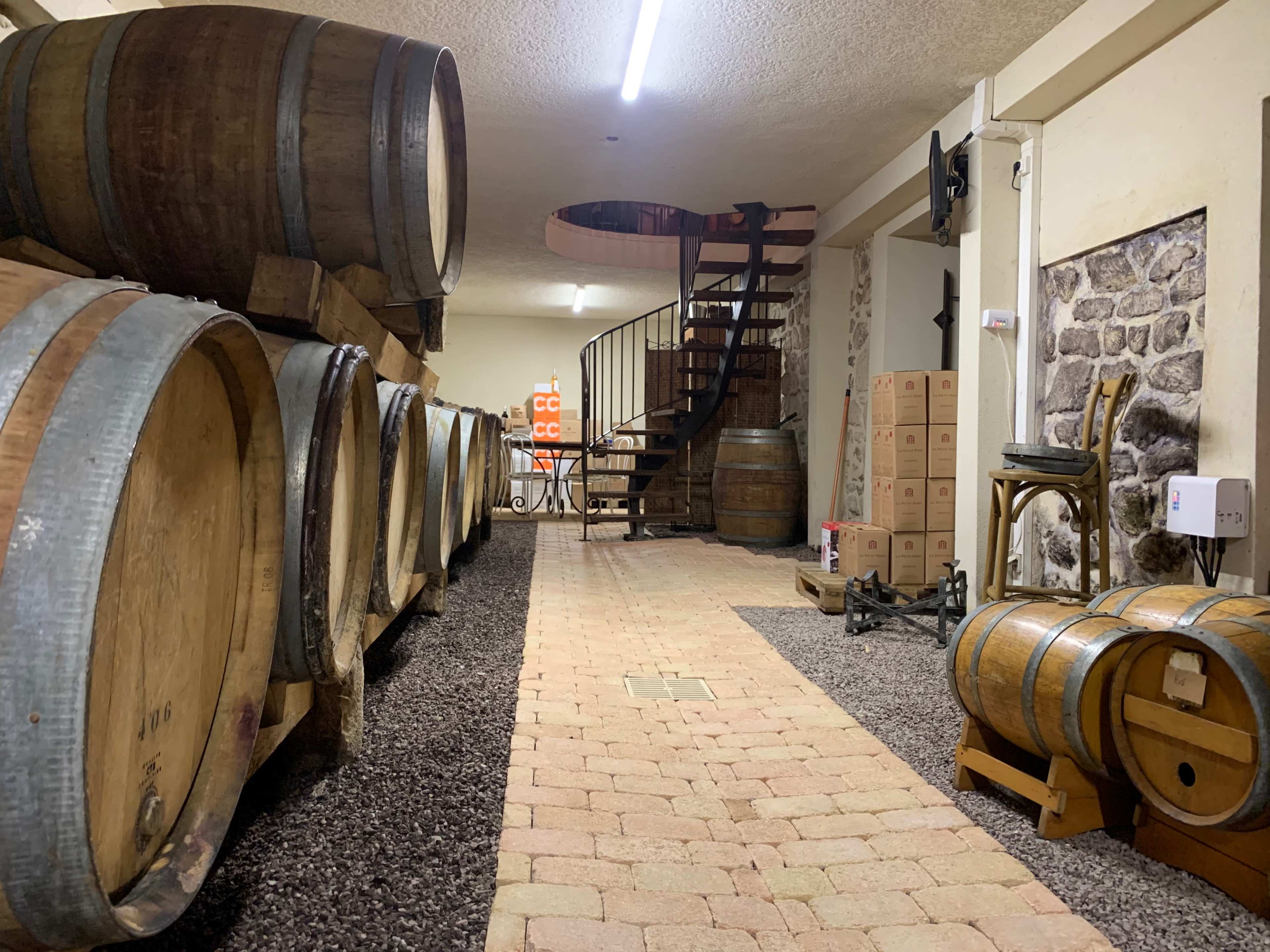 A cellar with large wooden barrels lined against the wall, a spiral staircase in the background, and boxes stacked on a table.