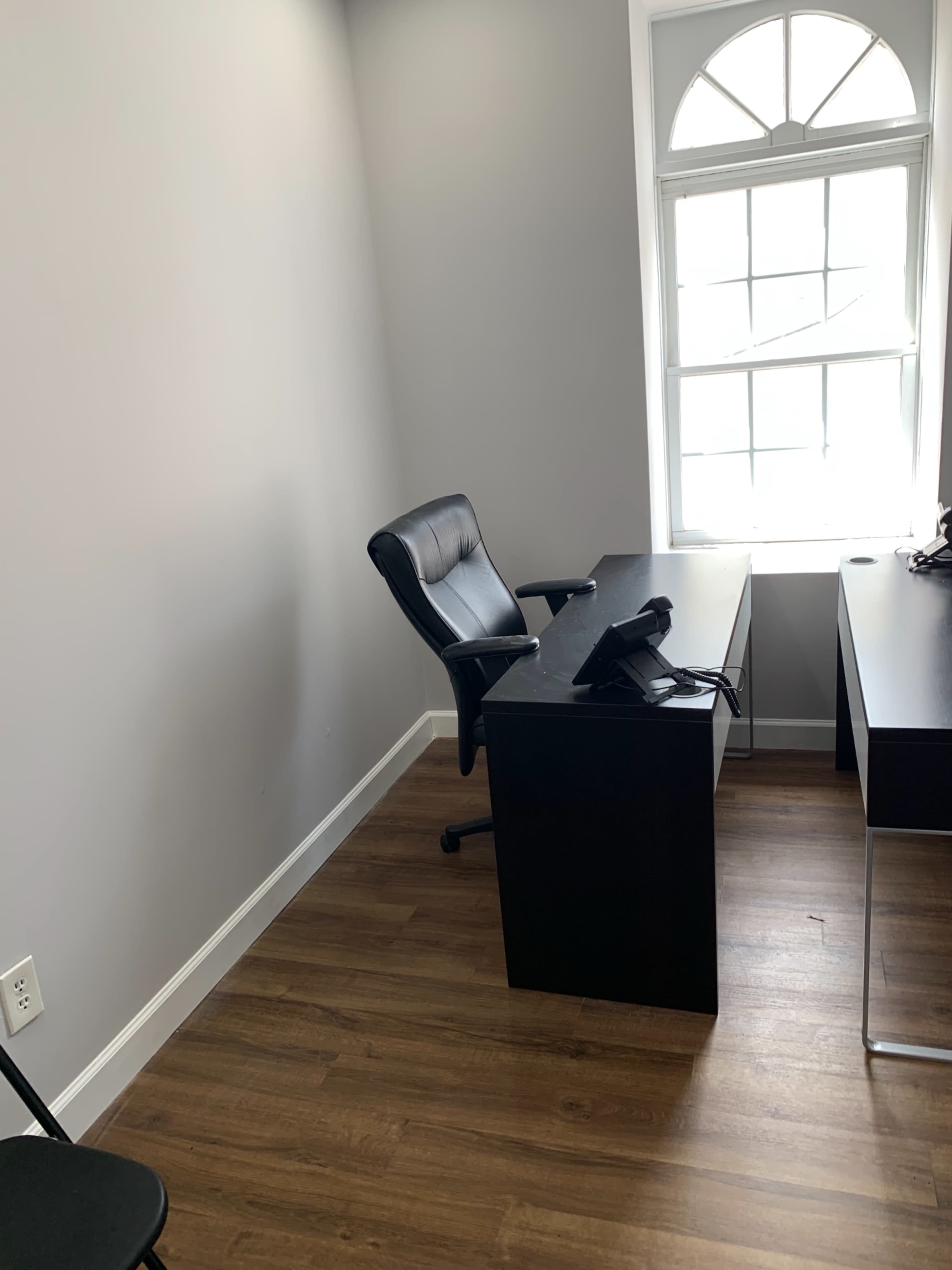 The image shows a small office space with a black desk, a black chair, and a window letting in natural light.