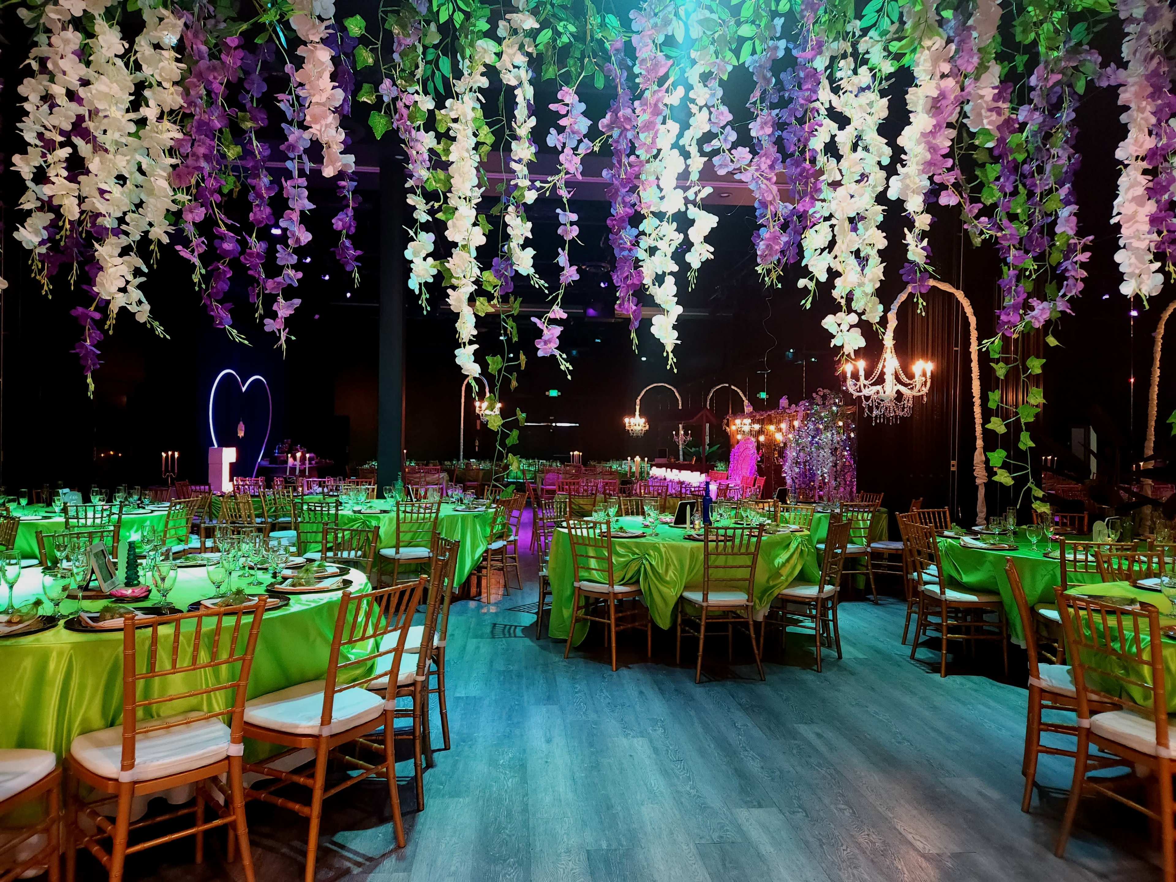 The image shows a banquet hall decorated with green tables and vibrant floral hangings, illuminated by colorful lights.