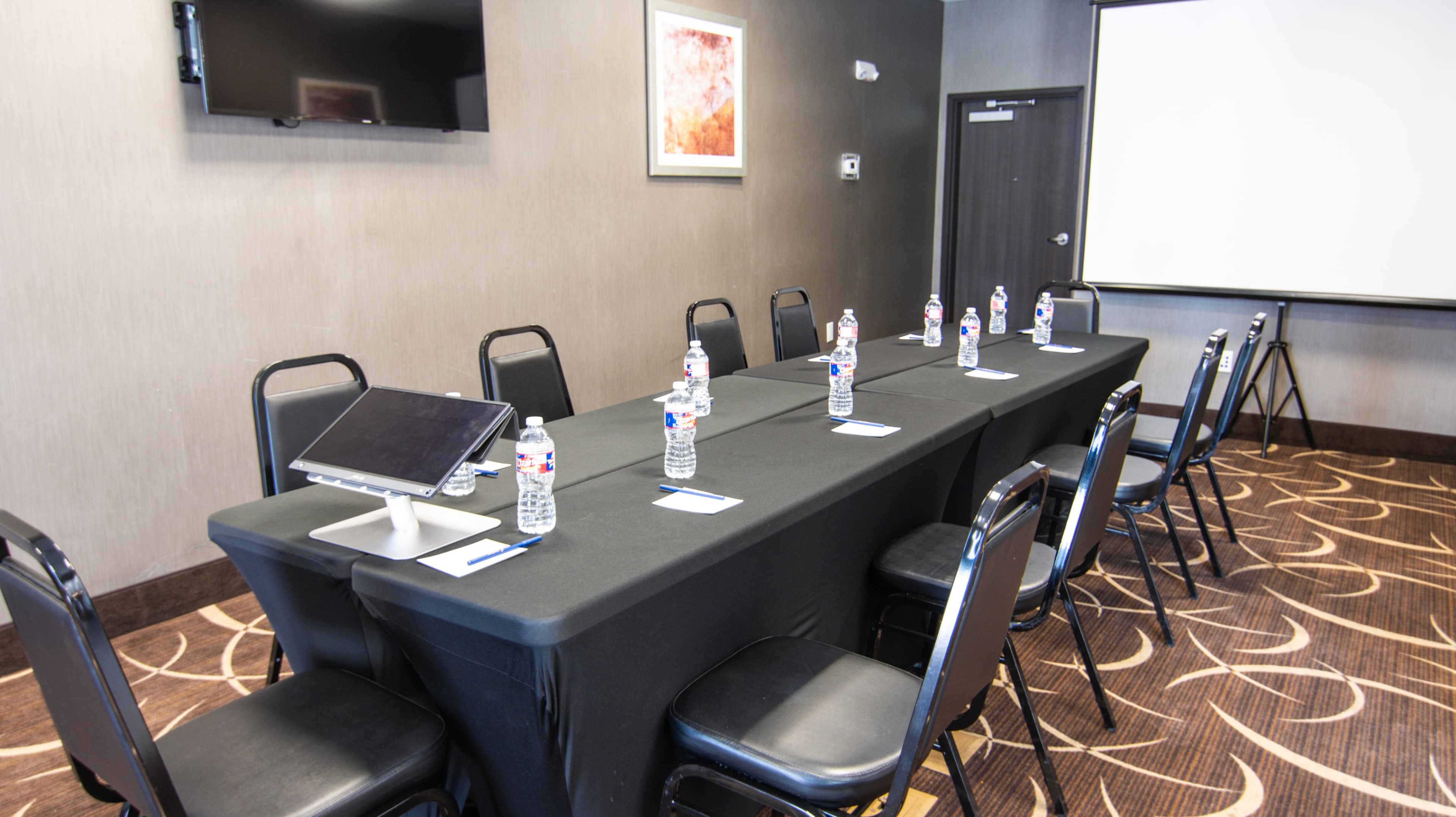 A conference room setup features a long table with chairs, each equipped with a bottle of water and a name tag, alongside a laptop and a screen for presentations.