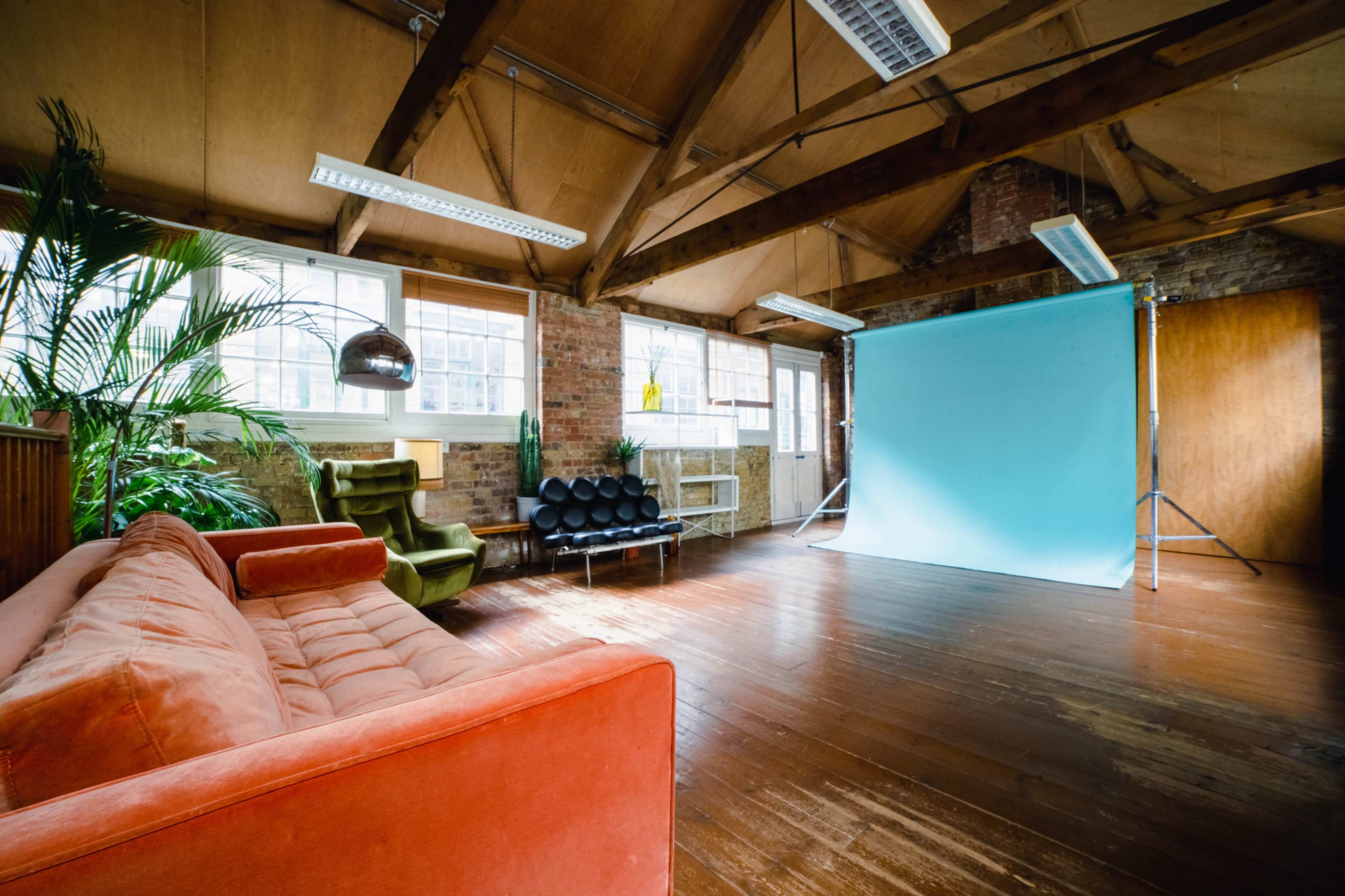 A spacious room with hardwood floors, large windows, and a blue backdrop set up for photography, featuring a mix of modern furniture including an orange sofa and black chairs.