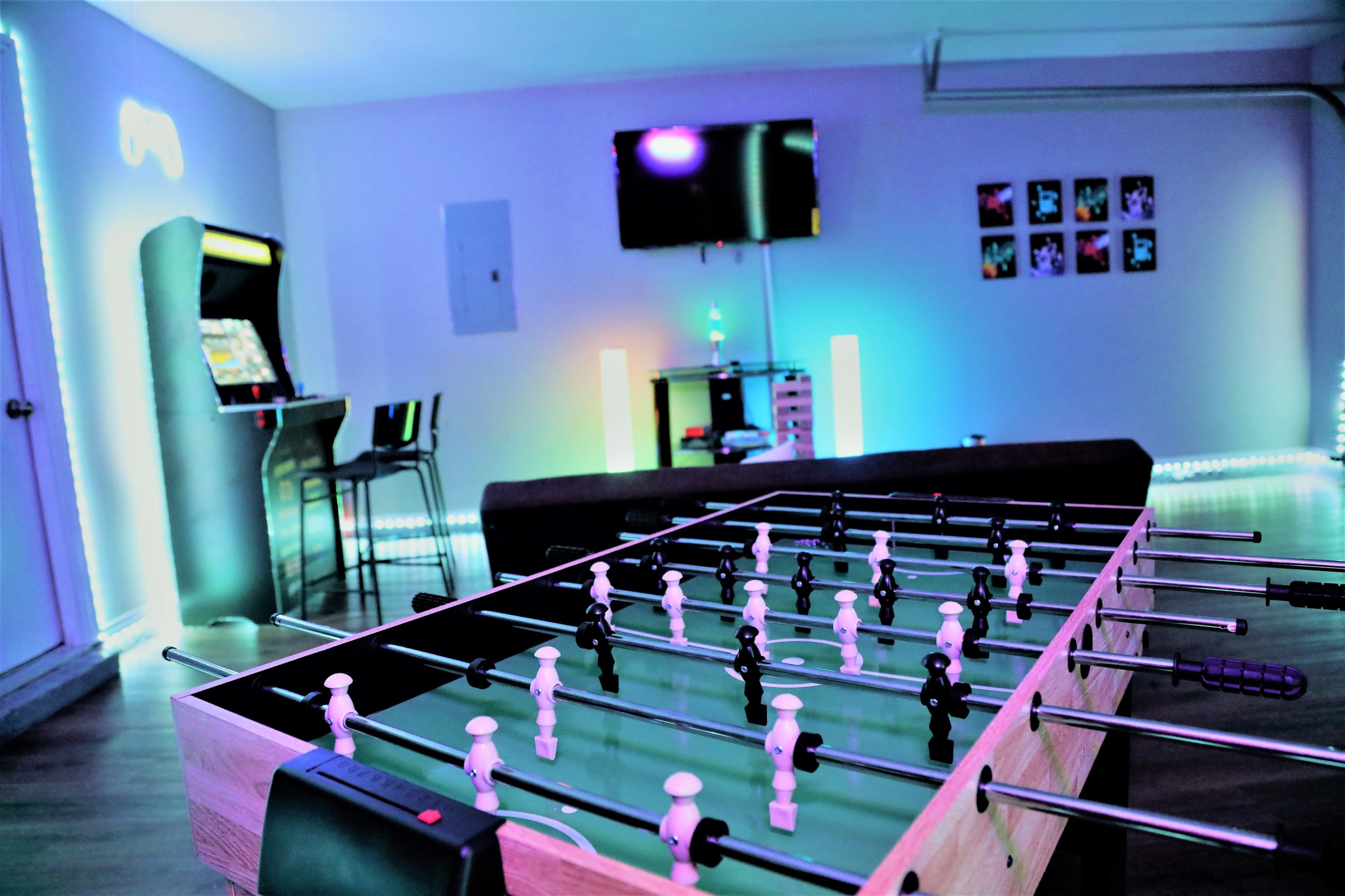 A foosball table in a brightly lit game room with an arcade machine, a television, and colorful wall art.