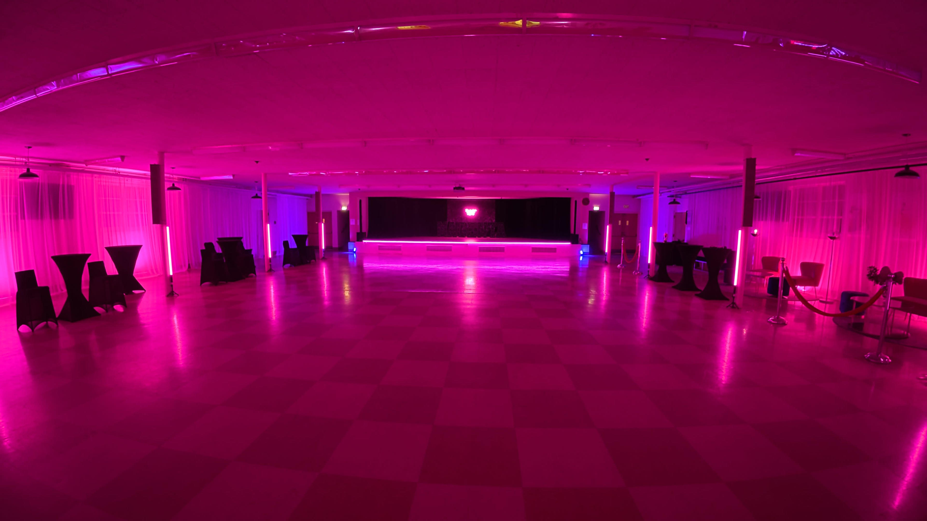 A spacious event hall is illuminated with pink lighting, featuring a checkered floor and a central stage area.
