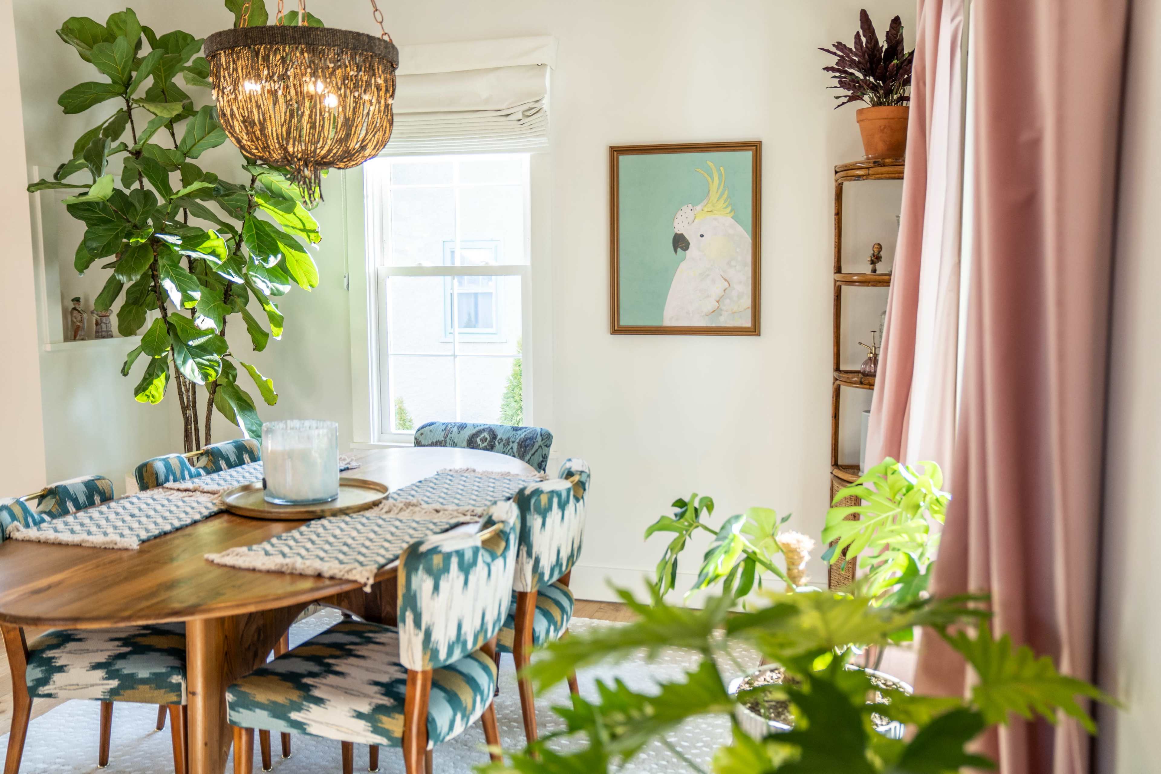 A dining area features a round wooden table surrounded by patterned chairs, with a large window, potted plants, and a framed artwork on the wall.