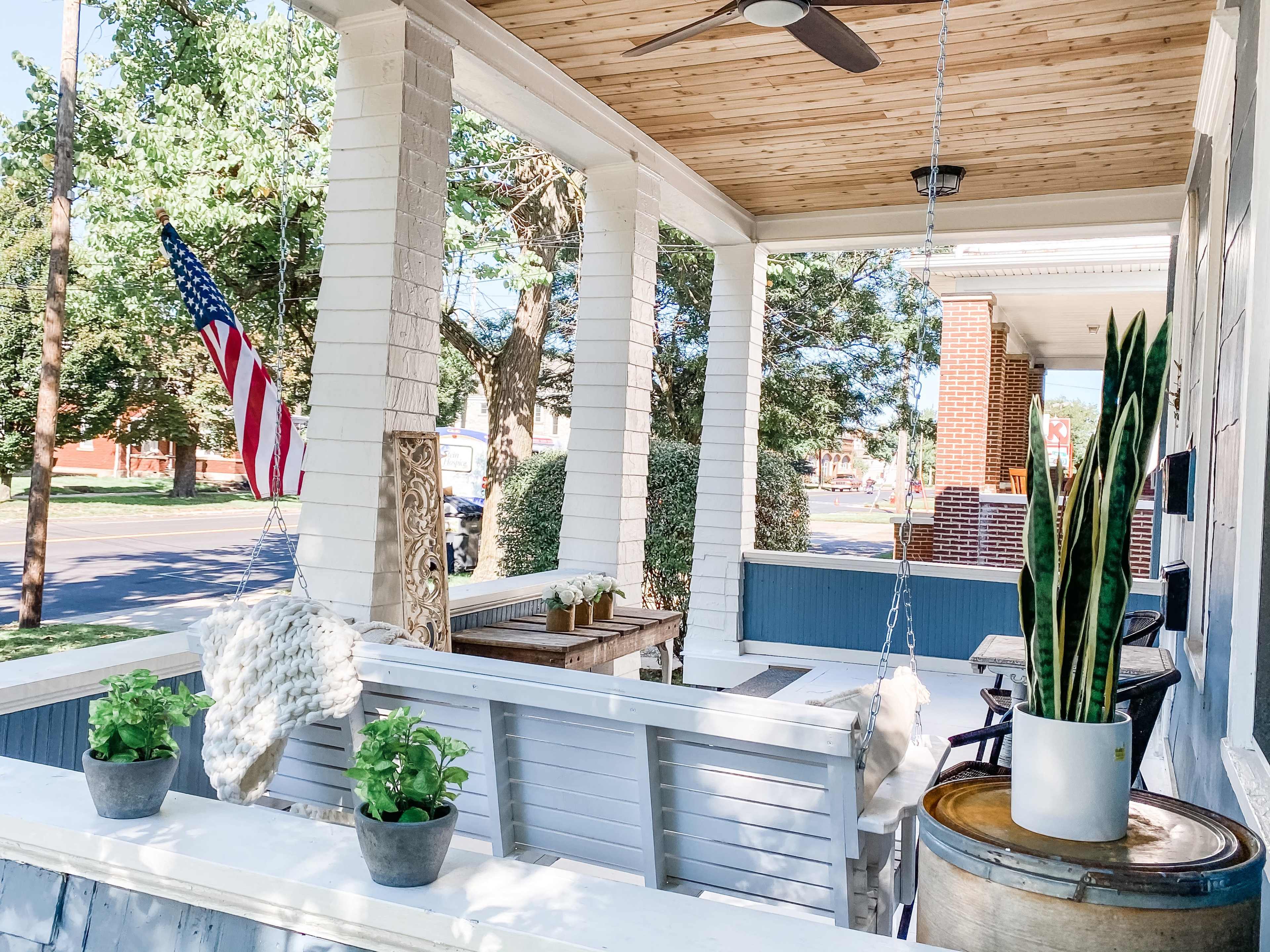 A front porch featuring a swing, potted plants, and an American flag, with trees and a street visible in the background.