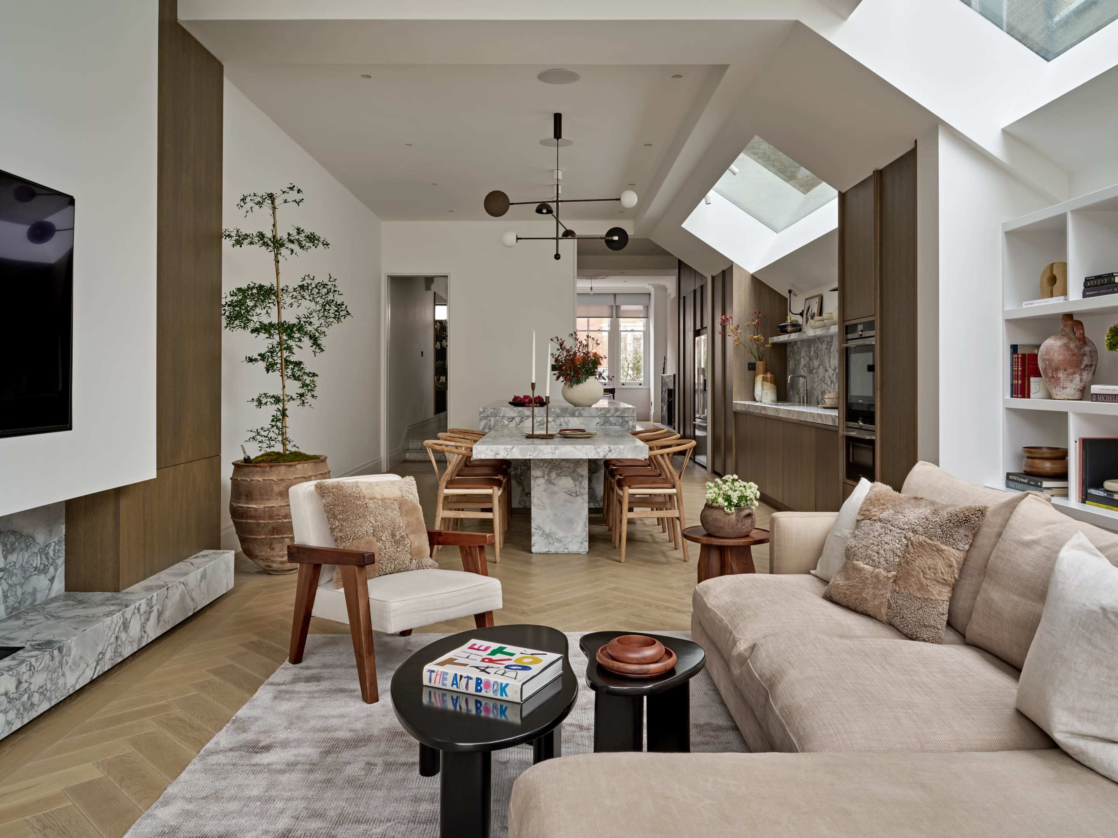 The image shows a modern open-concept living space featuring a light-colored sofa, a marble dining table, and large windows that allow natural light to illuminate the room.