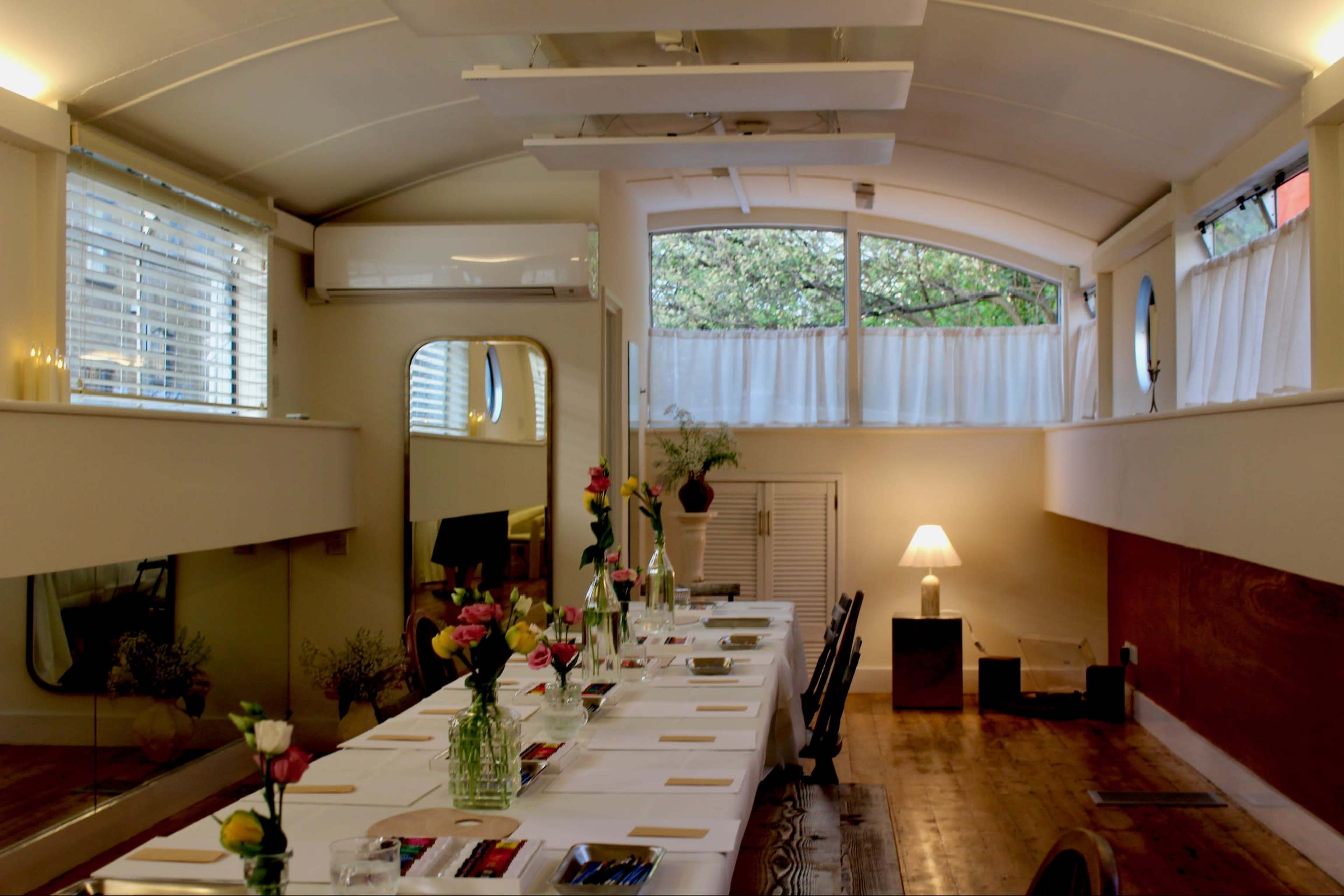 The image shows a well-lit interior space featuring a long dining table set with flowers and placemats, surrounded by minimalist decor and large windows.