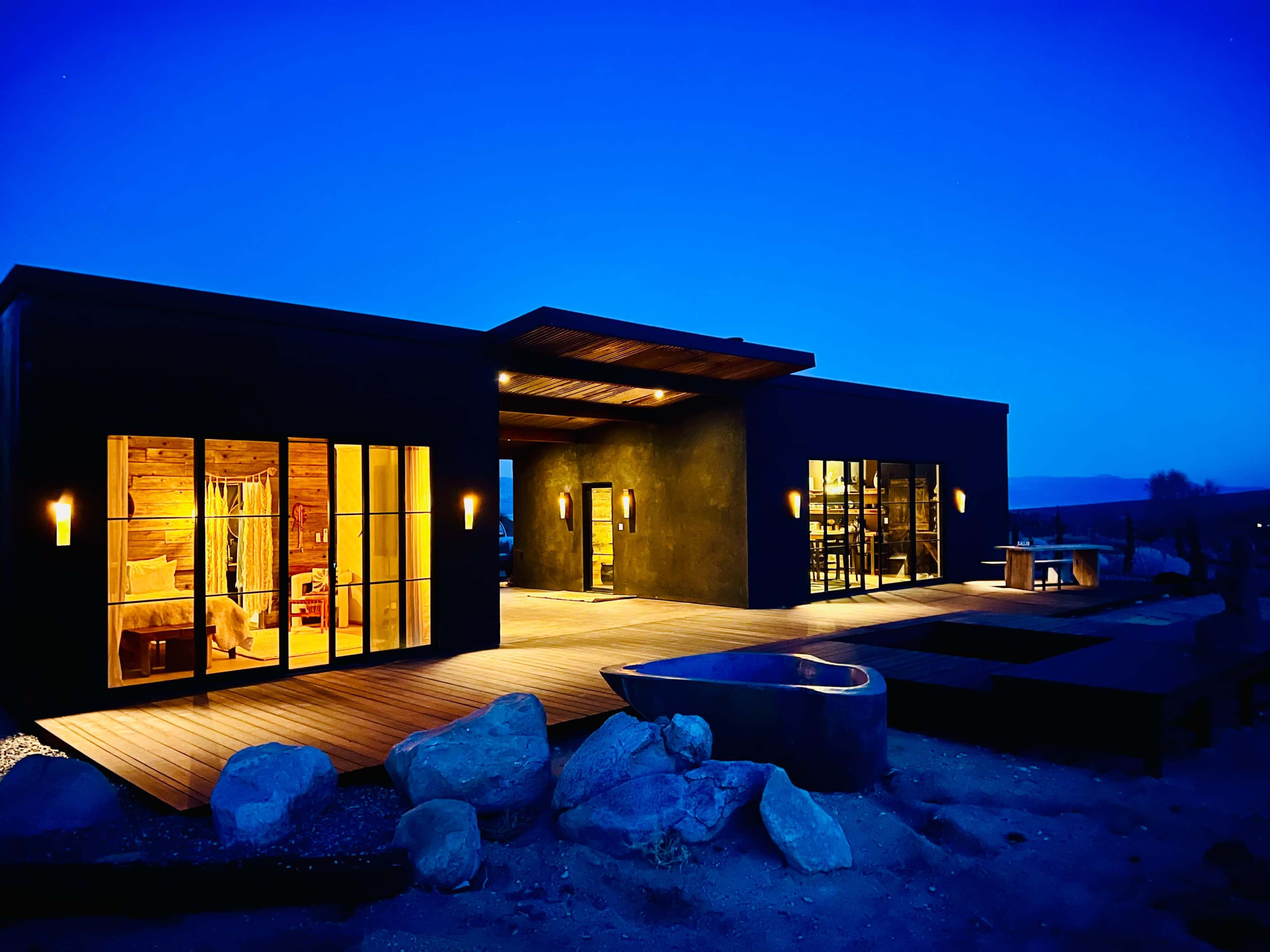 A contemporary house with large windows and a wooden deck is illuminated at dusk, surrounded by rocks and desert landscape.