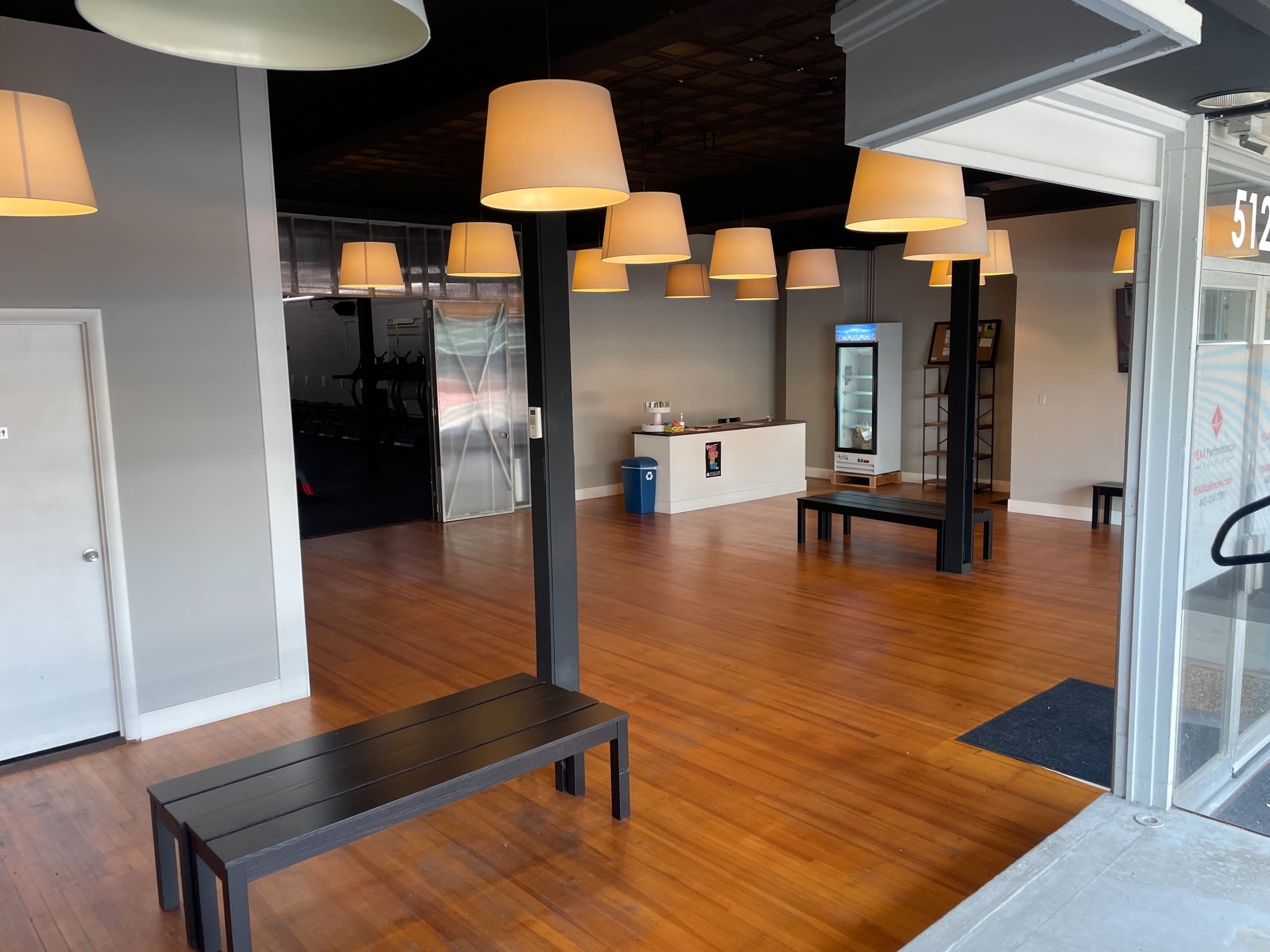 A spacious interior with wooden flooring and several pendant lights hanging from the ceiling, featuring benches along the sides and a service area in the back with a refrigerator.