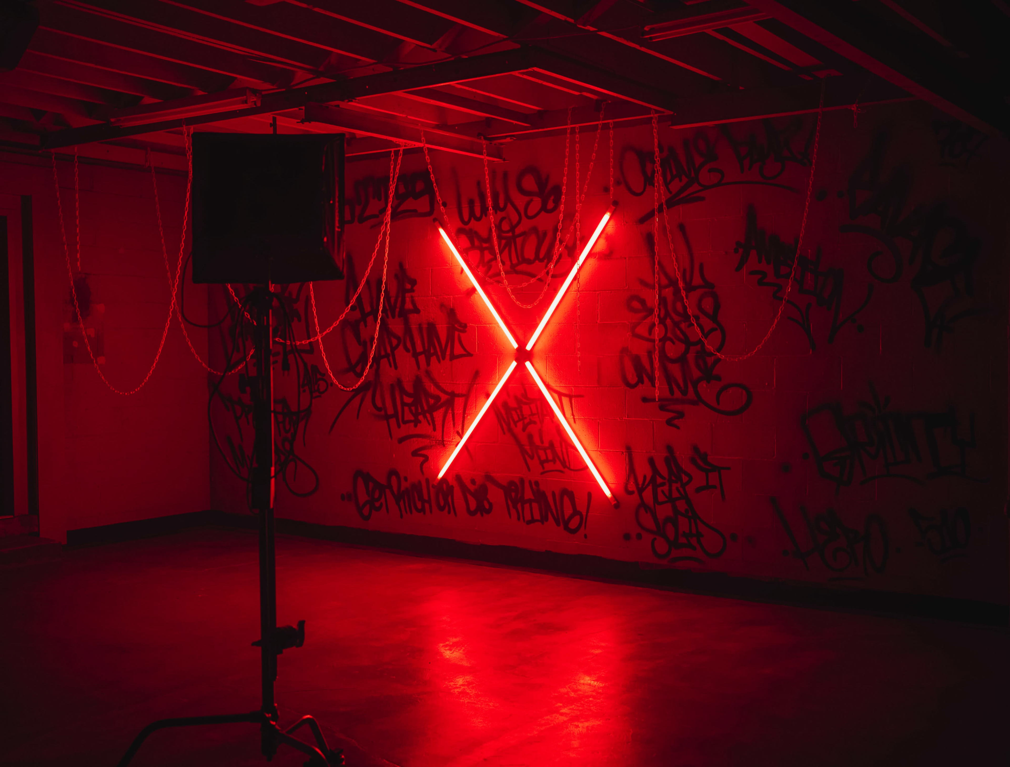 A dimly lit room features an illuminated red X made of neon lights against a wall covered in vibrant graffiti.