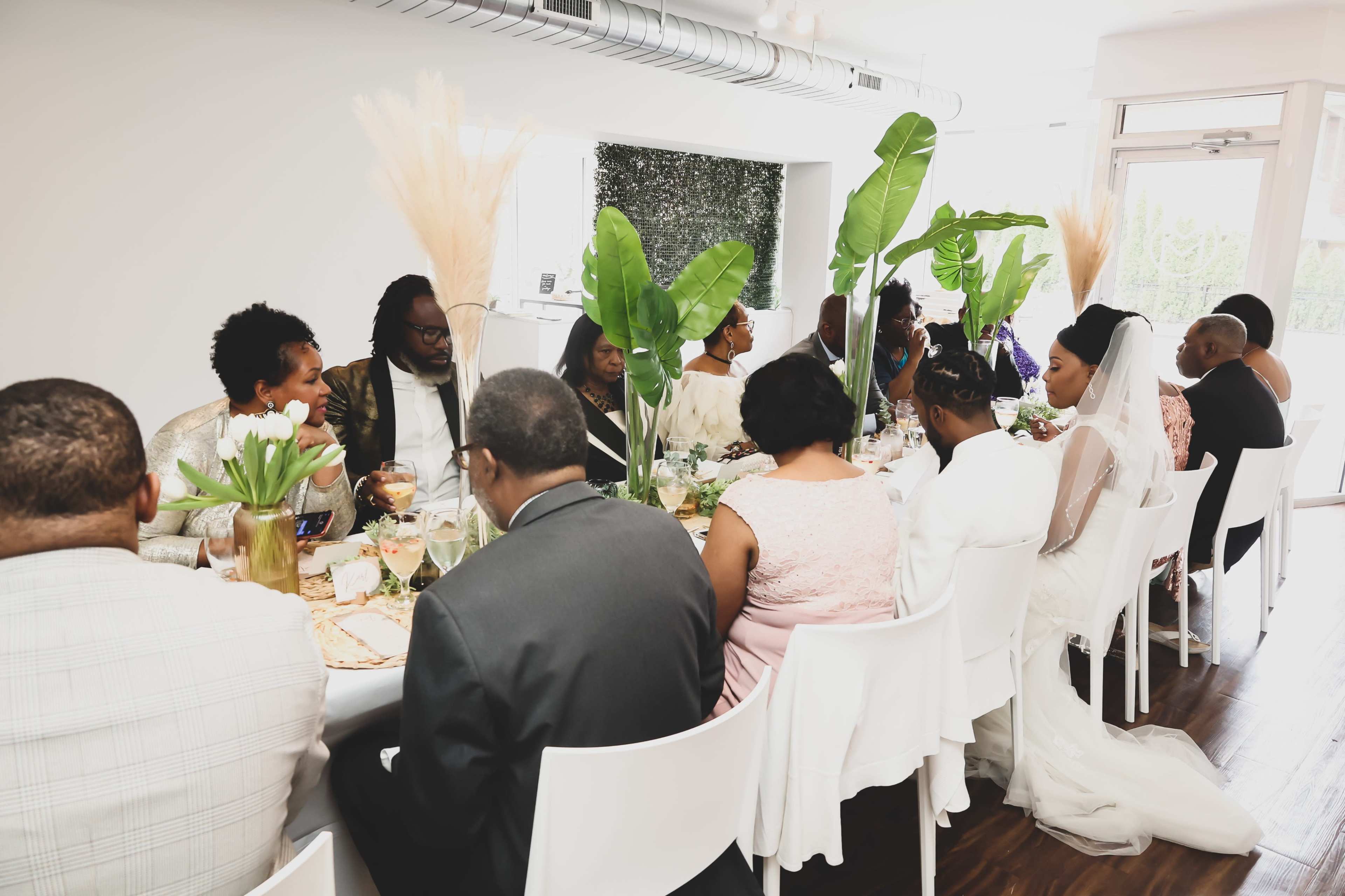 A group of people sit at a long dining table adorned with greenery and decorative centerpieces in a bright, modern venue.