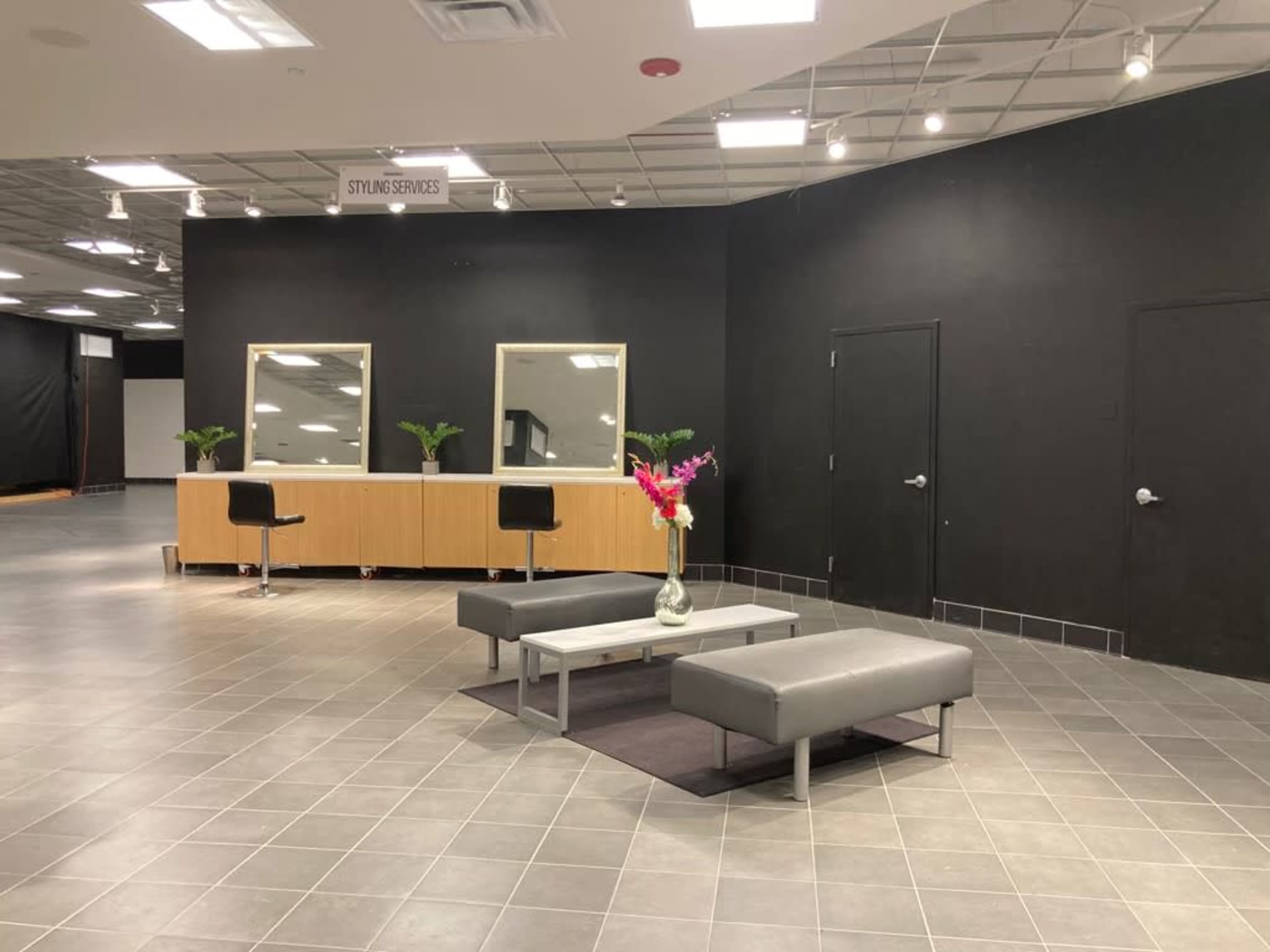 The image shows a modern interior space with a seating area featuring a table and gray couches, flanked by a reception area with three mirrors and a counter against a black wall.
