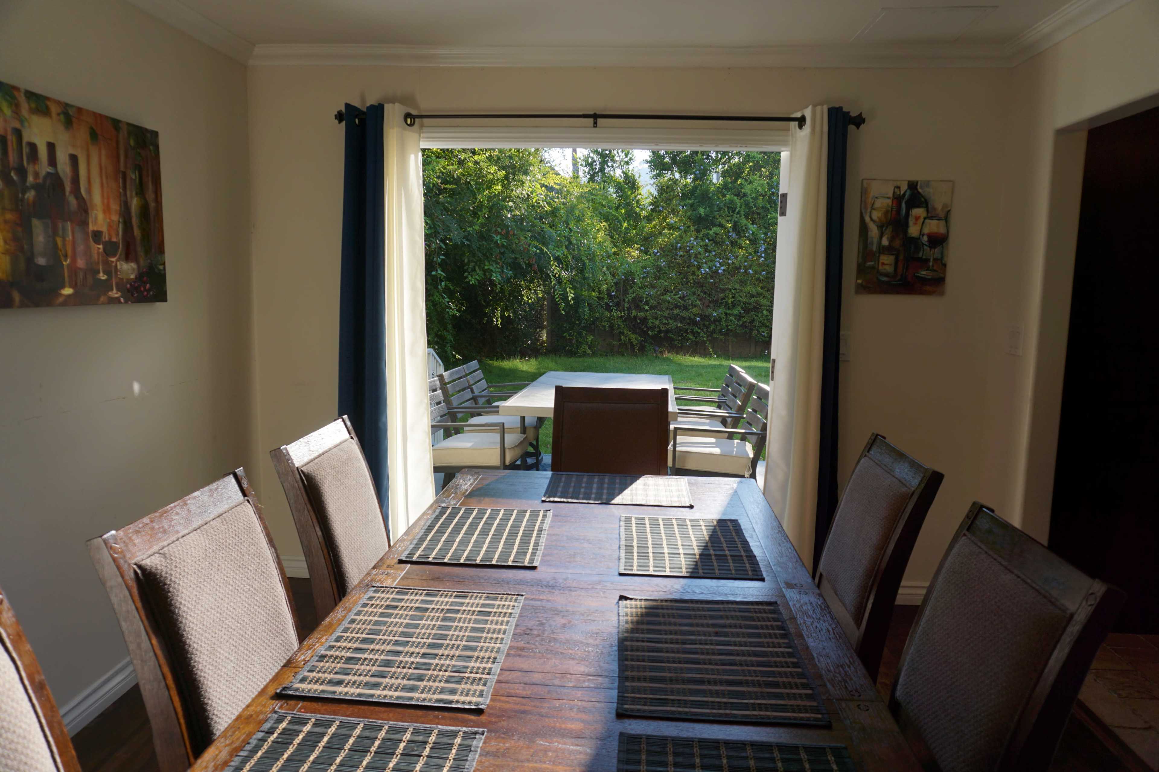 A spacious dining area features a long wooden table with woven placemats, looking out through an open sliding door to a patio with outdoor furniture and a green landscape.
