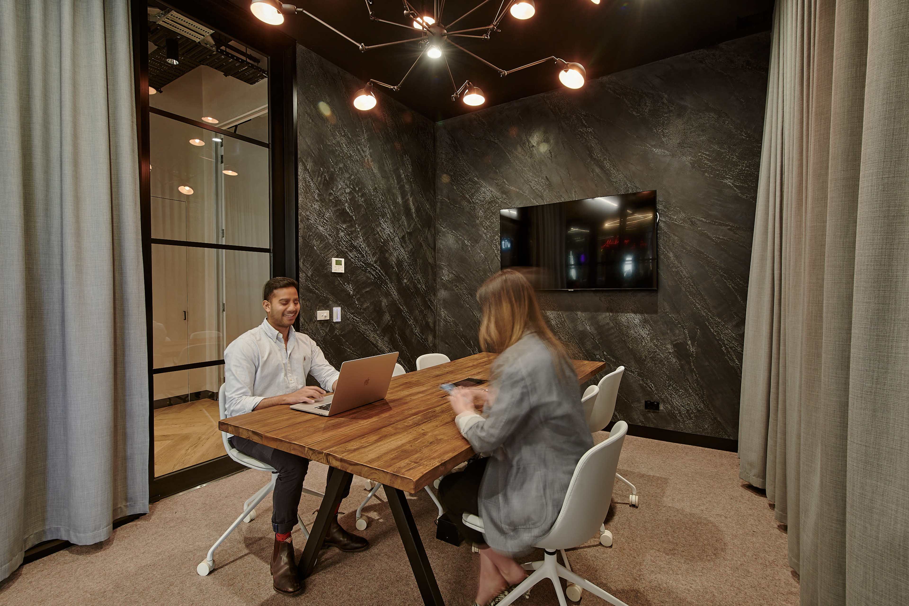 A meeting room features a wooden table with two people seated, one using a laptop while the other looks at a mobile device, surrounded by dark walls and modern lighting fixtures.