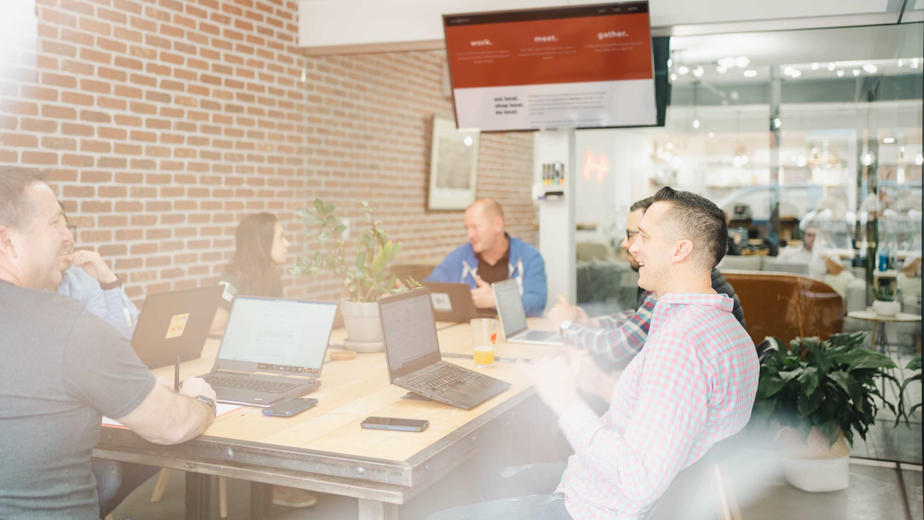 A group of people are sitting around a wooden table in a meeting room, engaged in discussion with laptops open in front of them.