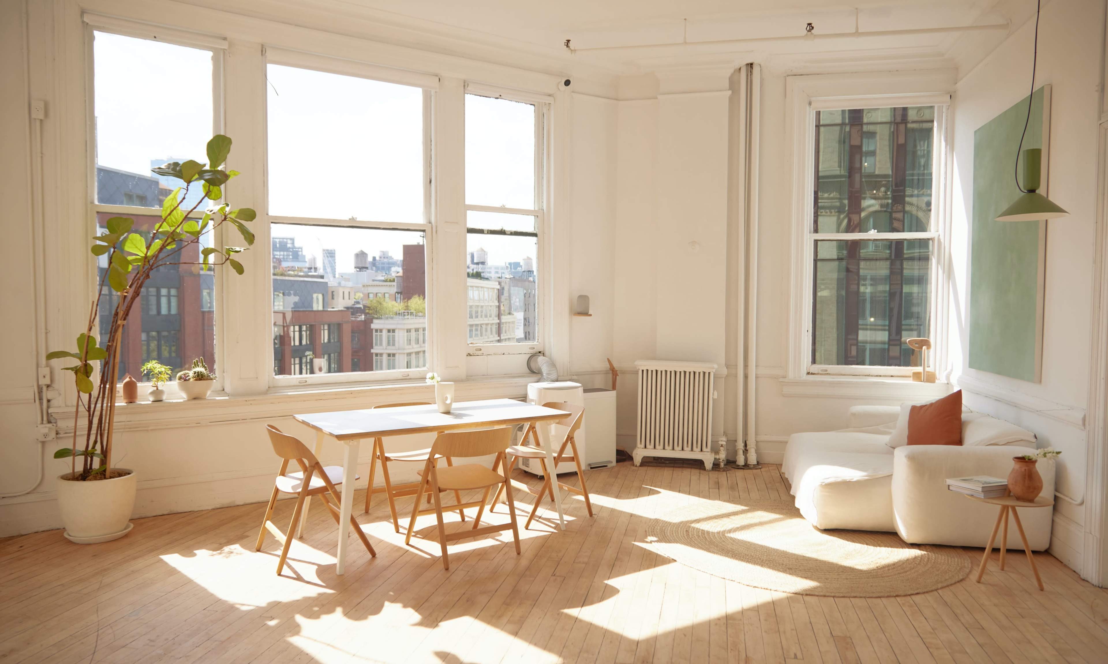 A bright, airy room with large windows, a wooden dining table with chairs, and a light-colored sofa, all set on a wooden floor.