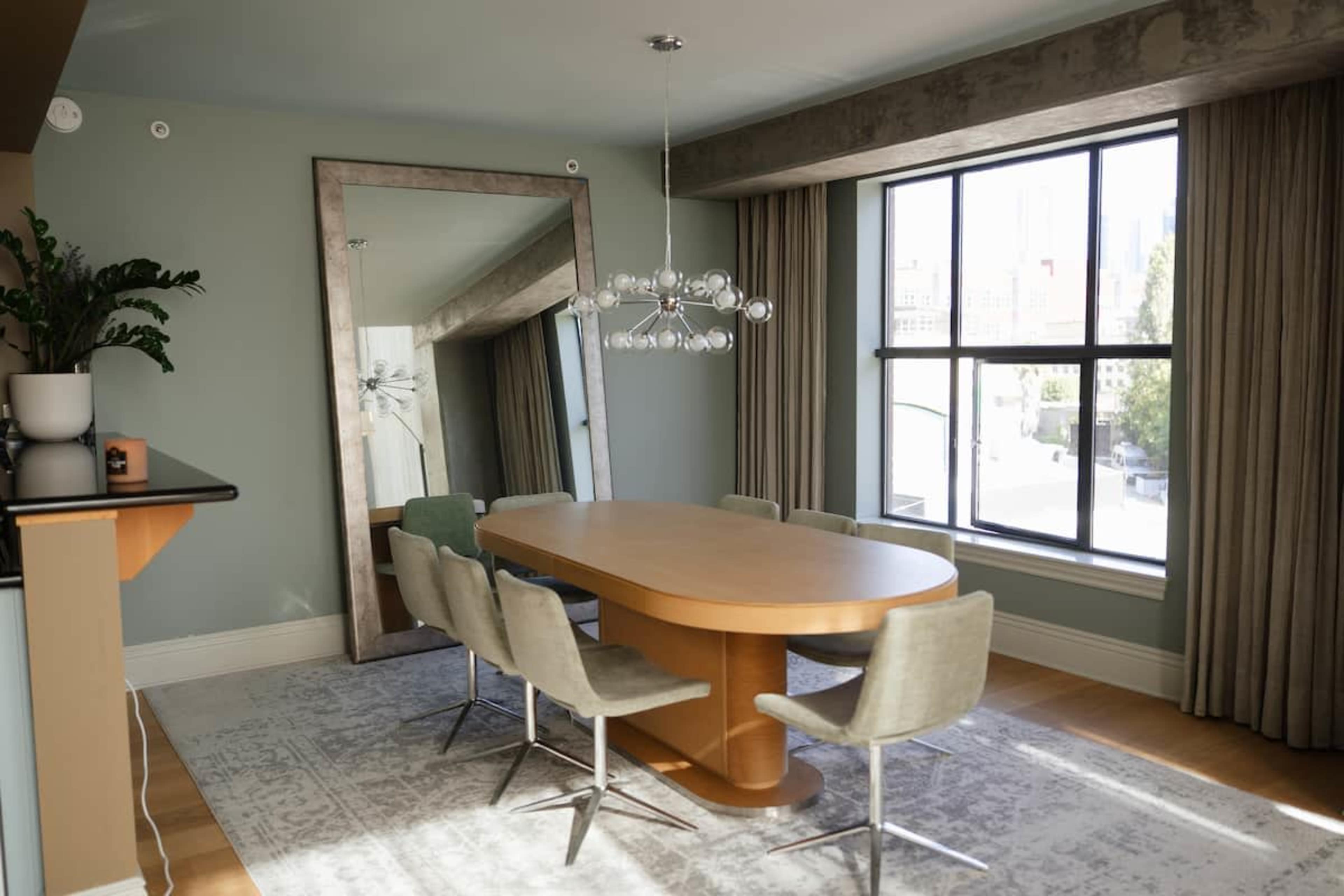 A modern dining room features an oval wooden table surrounded by gray chairs, with a large mirror and a unique light fixture, all set under a window with city views.