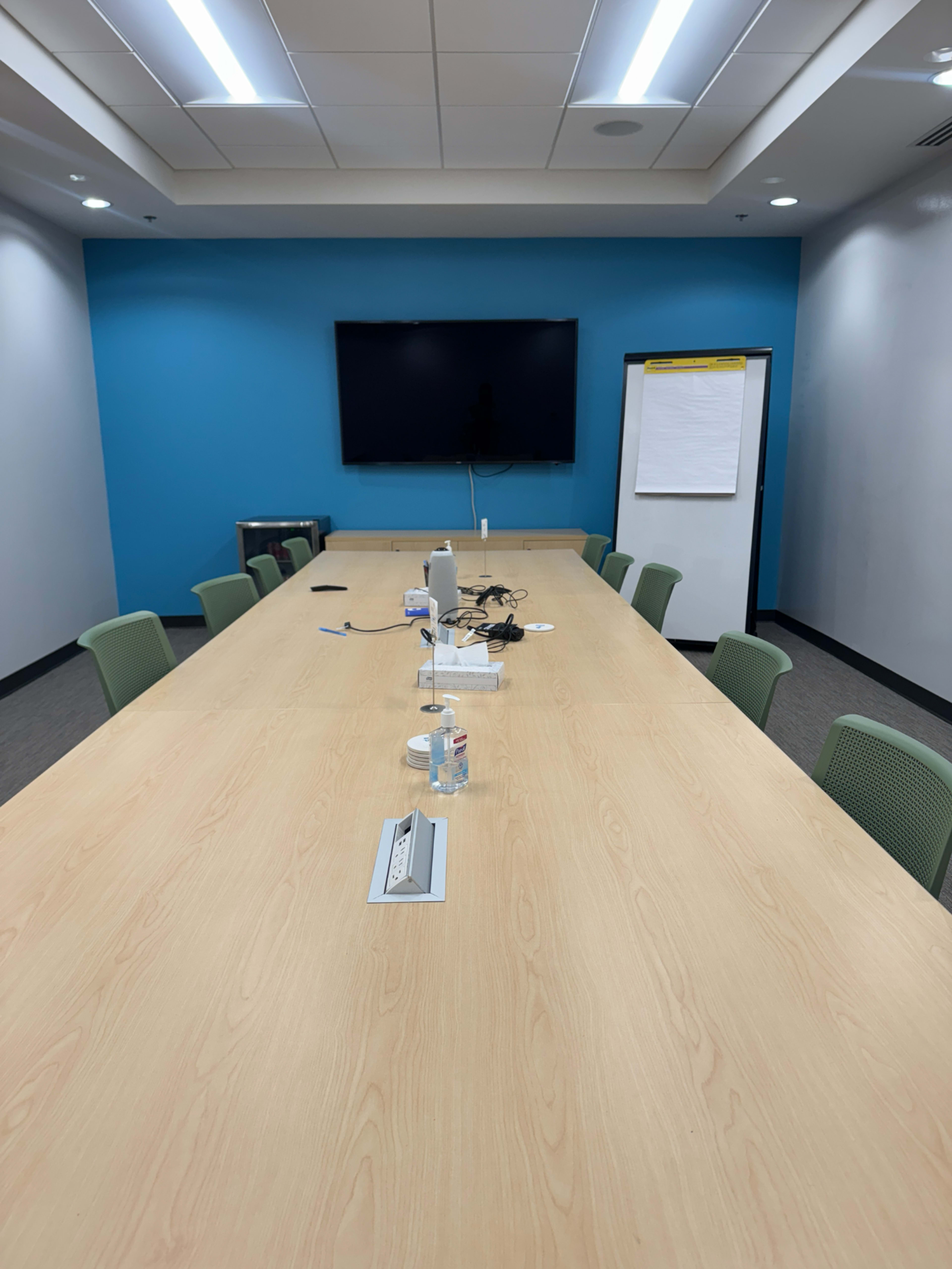 A long wooden conference table is set up in a meeting room with chairs around it, accompanied by a television on the wall and a flip chart nearby.