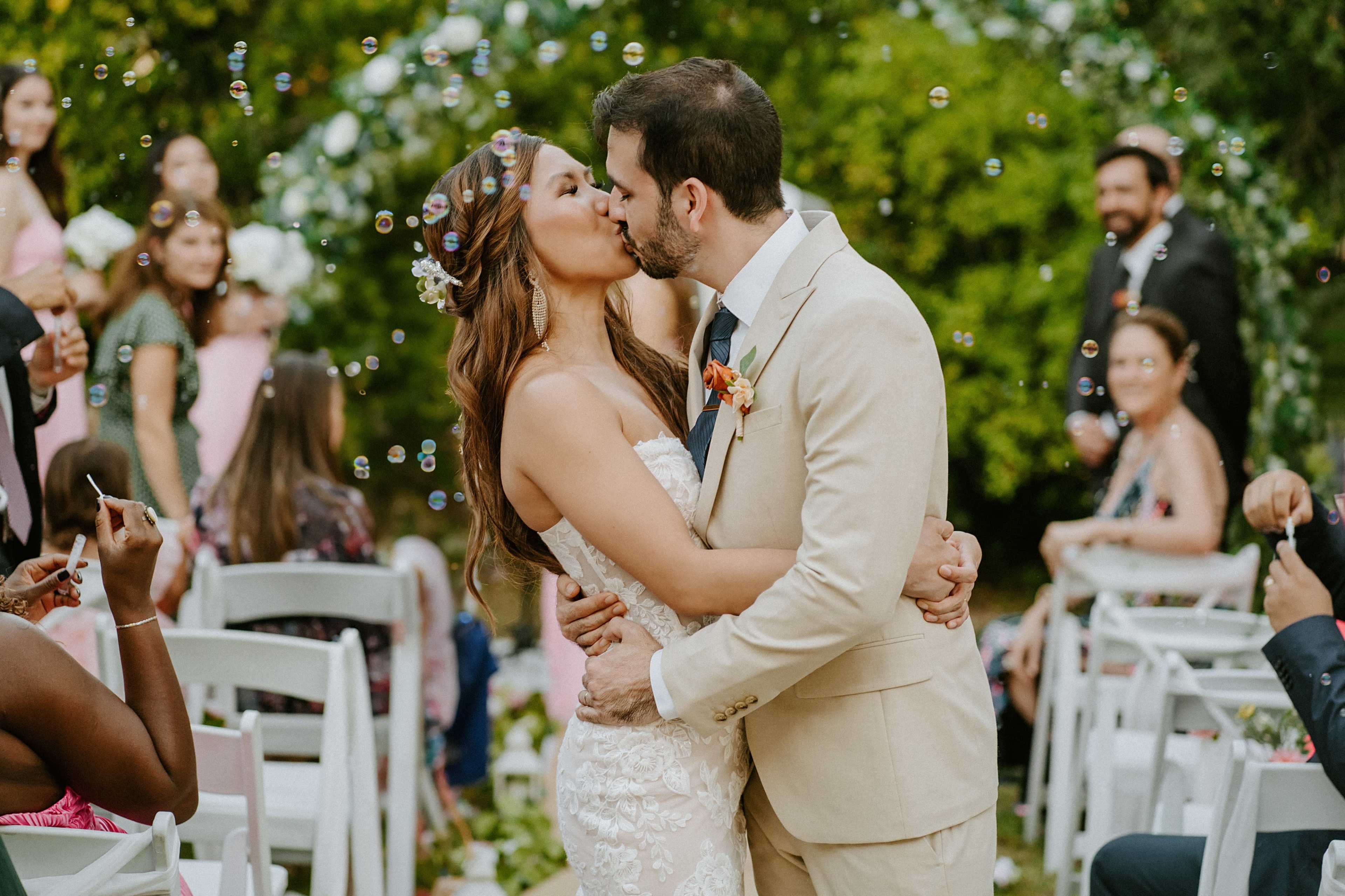 A couple exchanges a kiss while surrounded by guests celebrating at an outdoor wedding, with bubbles floating in the air.