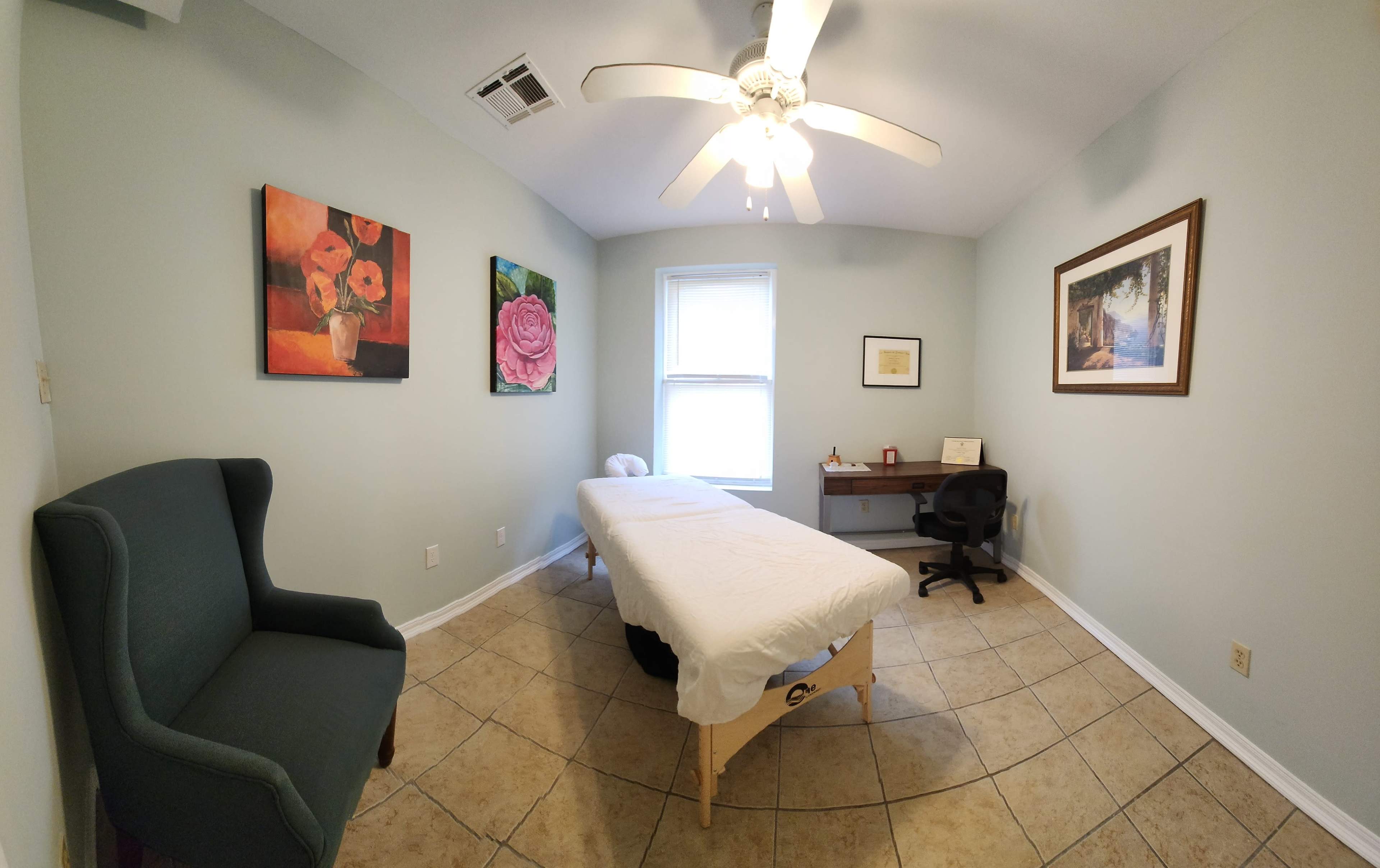 A treatment room with a massage table, a desk and chair, and two framed paintings on the walls.