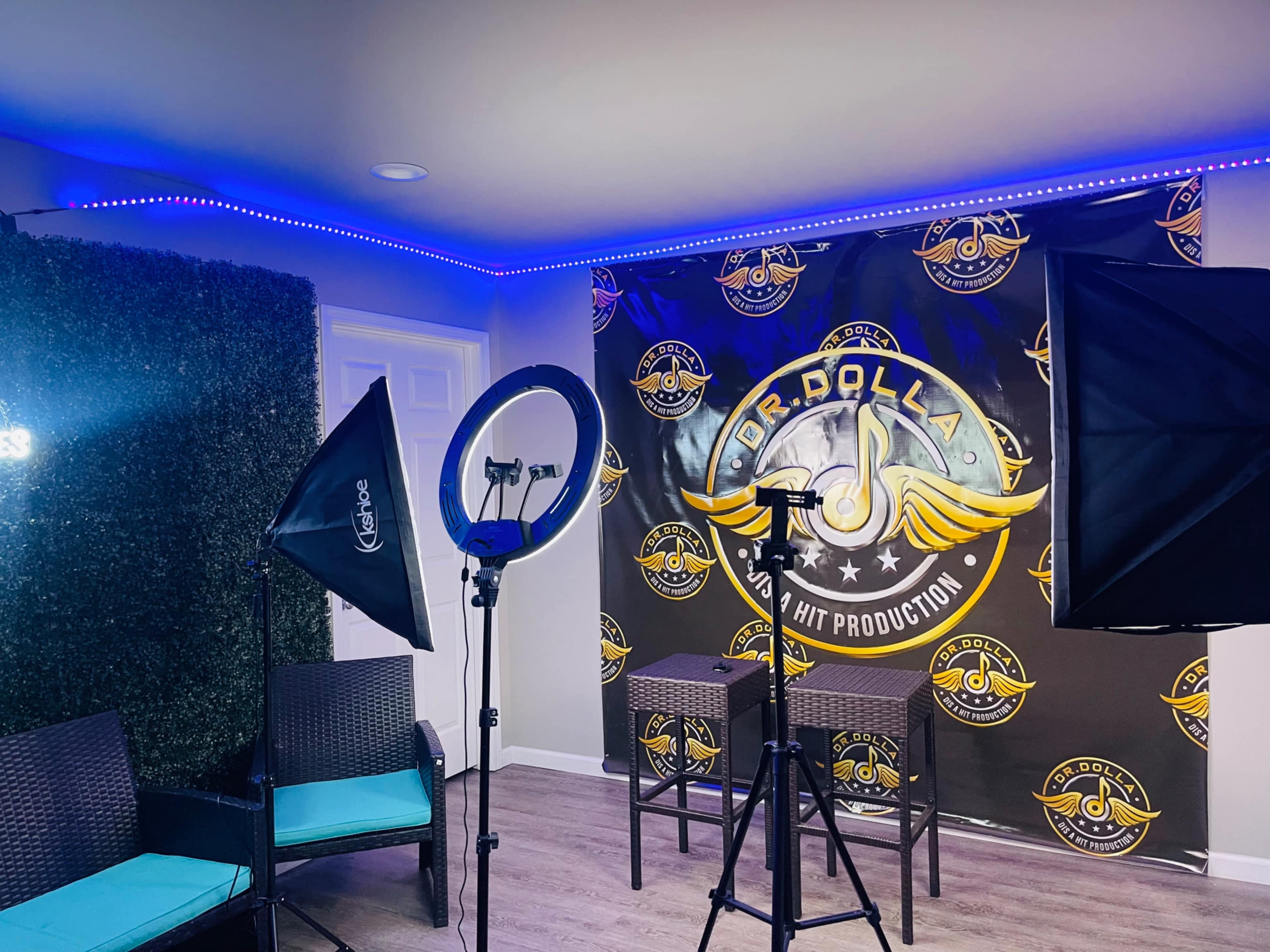 The image shows a recording studio setup with lighting equipment, two chairs, and a branded backdrop featuring a logo.