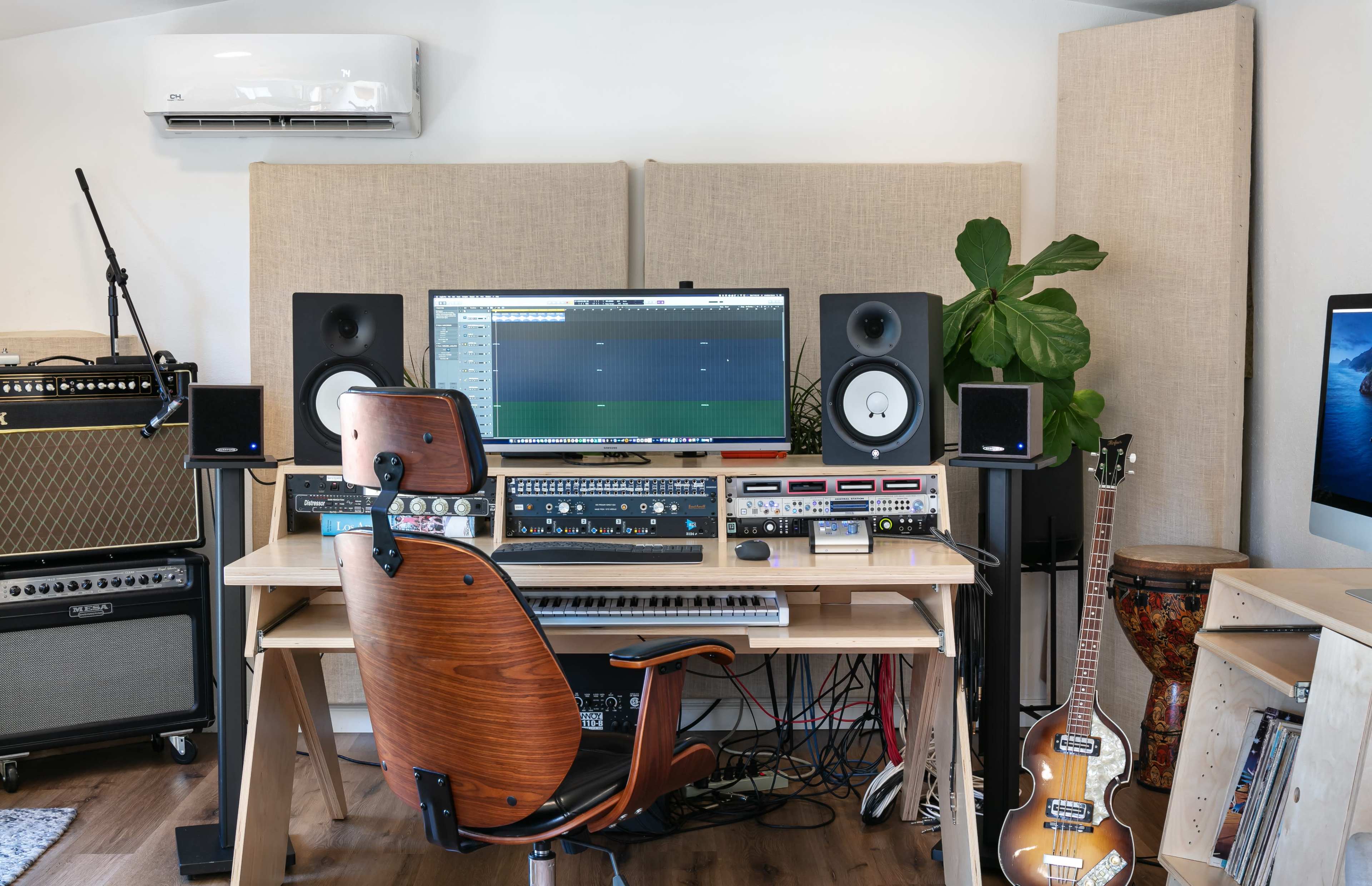 A modern music studio features a desktop computer, audio equipment, studio monitors, a keyboard, and various instruments, all set up in a clean workspace.