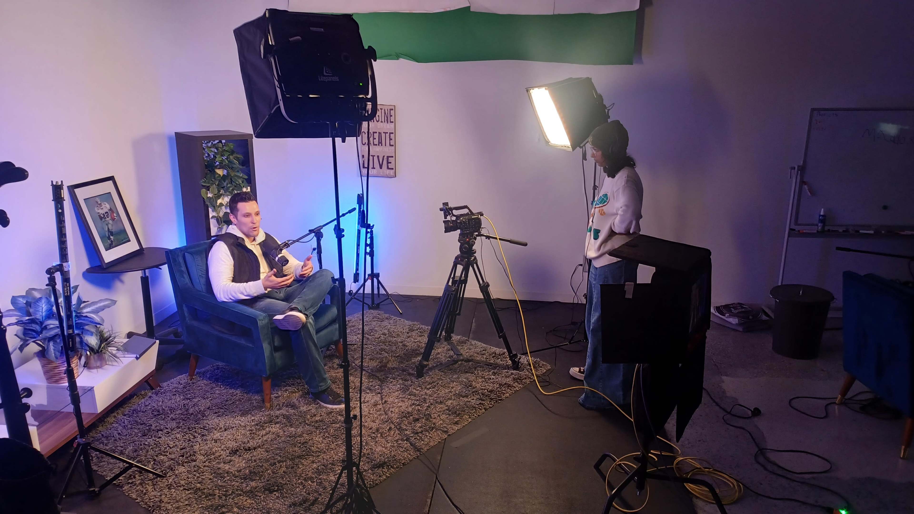 A man sits in a blue chair while a person stands beside a camera in a well-lit studio setup.