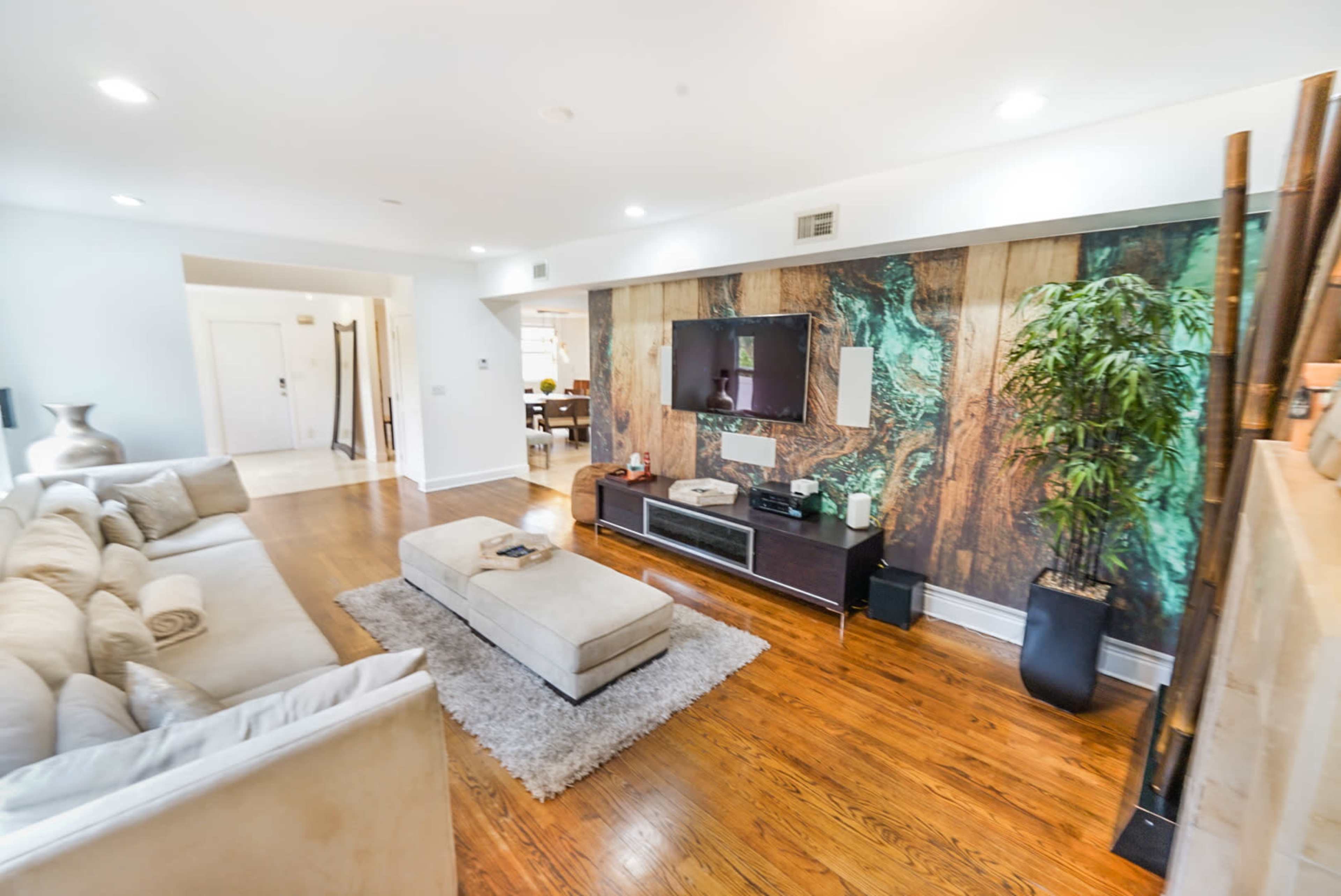 A spacious living room features a beige sectional sofa, a light-colored coffee table, a wall-mounted television, and a decorative mural amidst wooden accents.