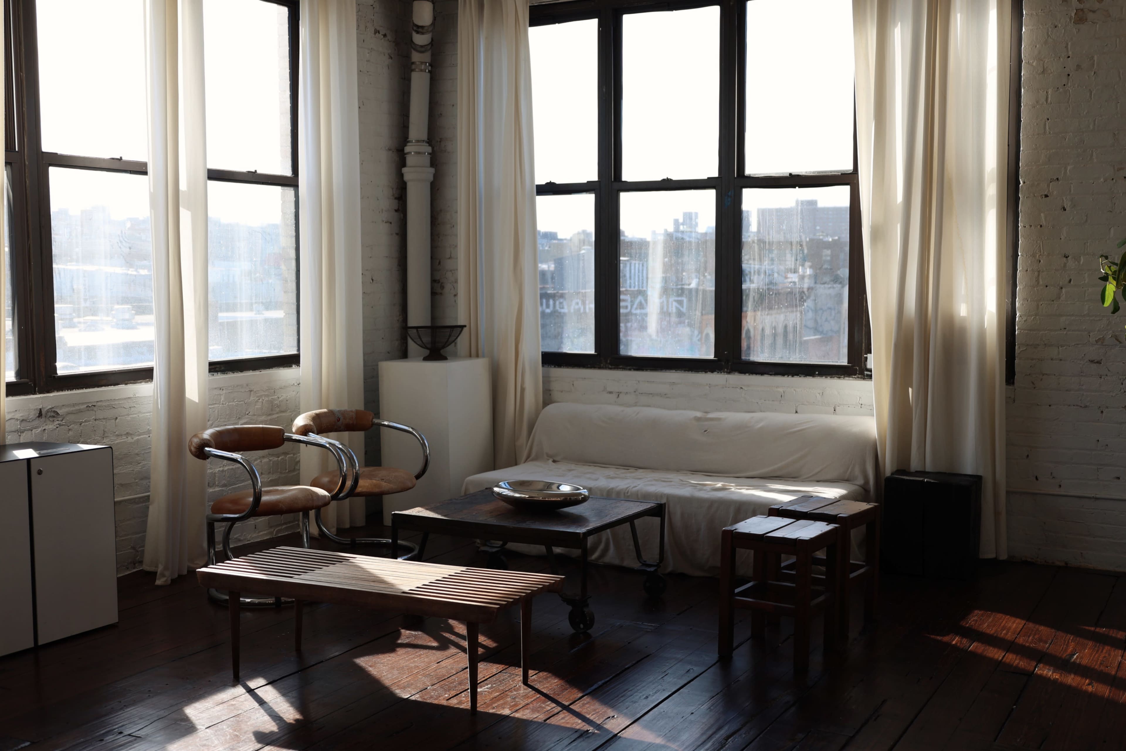 A spacious room features large windows with sheer curtains, showcasing minimal furniture including a sofa, two chairs, and a wooden coffee table.