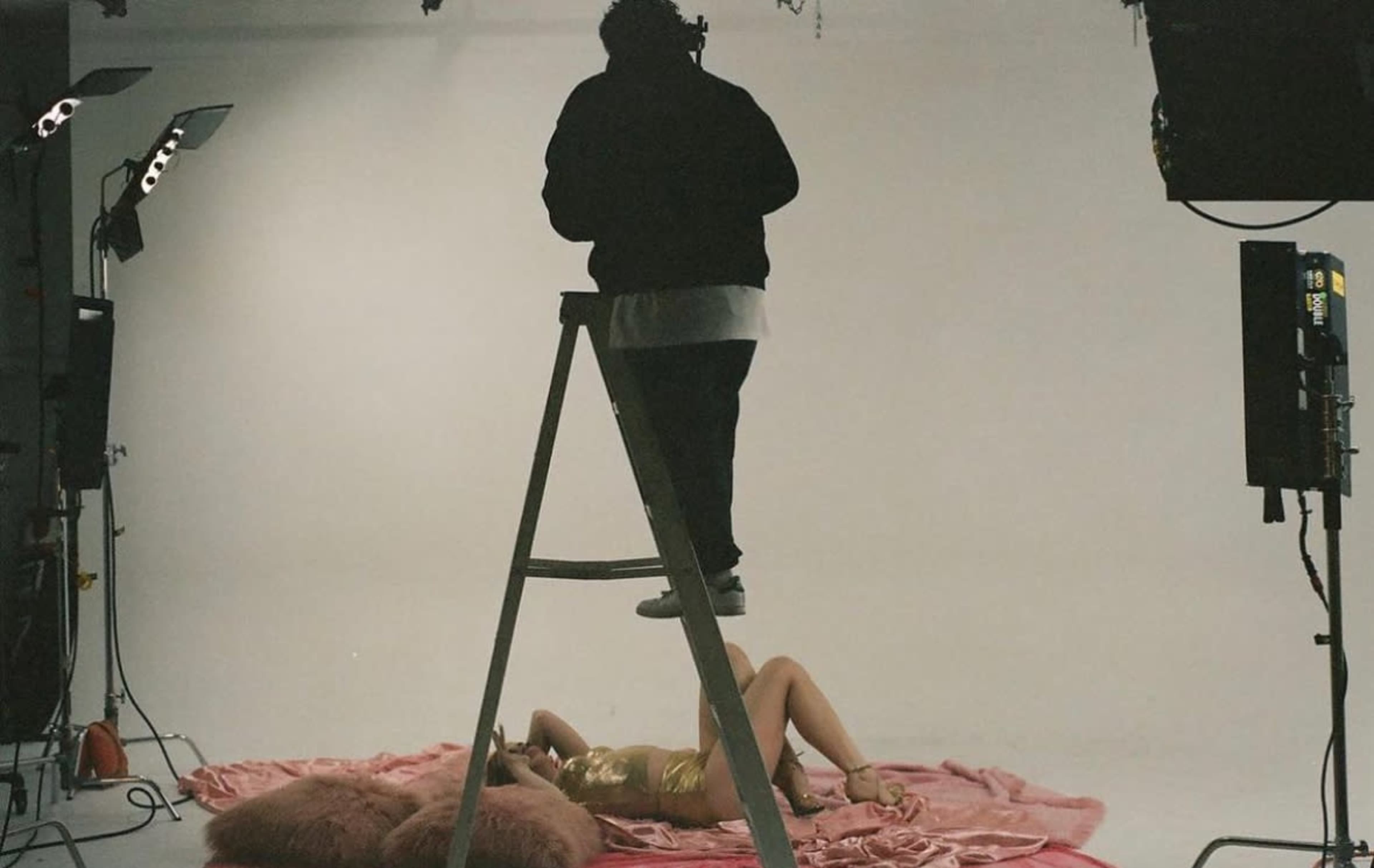 A person stands on a ladder above a model lying on a pink fabric backdrop in a photography studio.