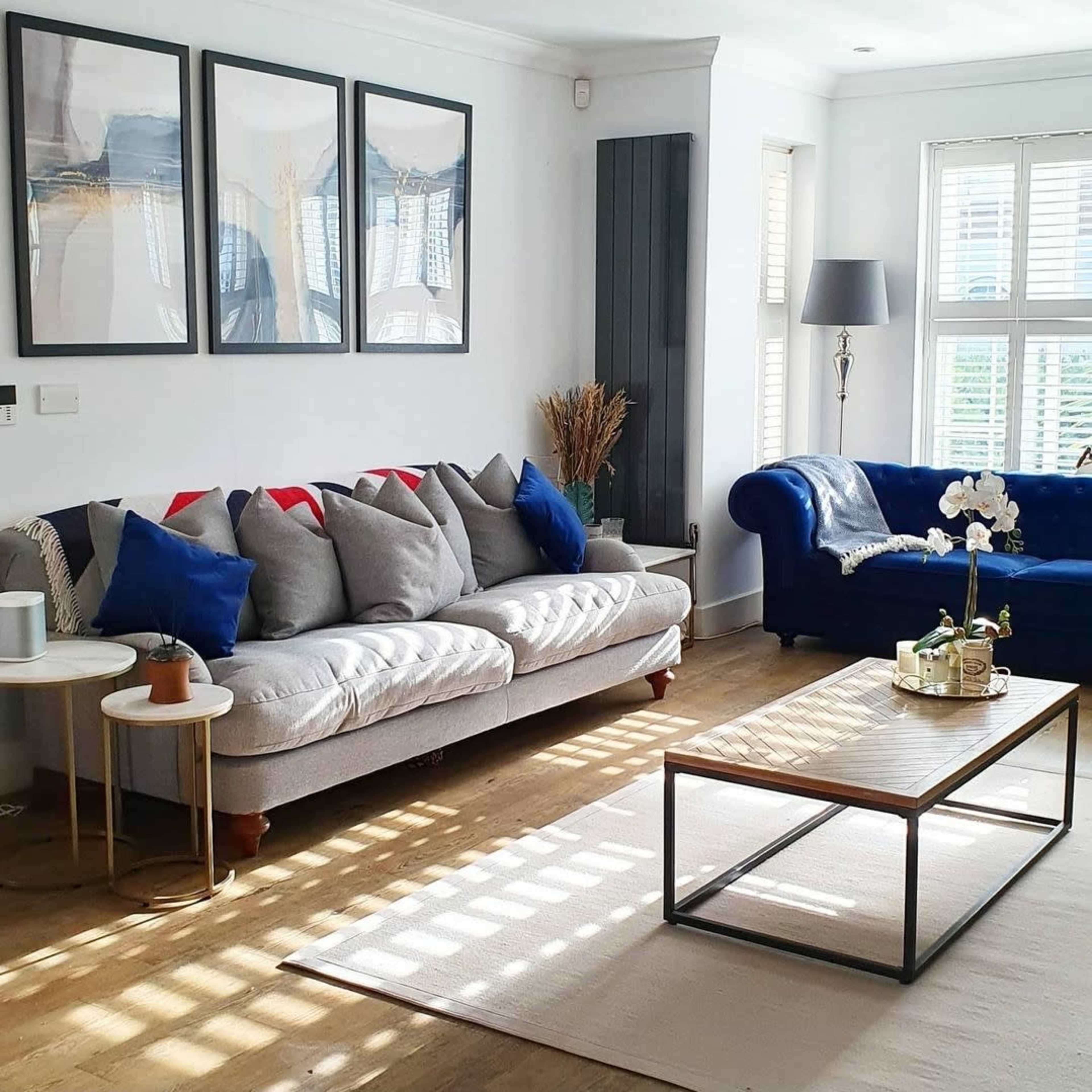 A living room features a gray sofa with colorful cushions, a blue chair, a wooden coffee table, and framed artwork on the walls.