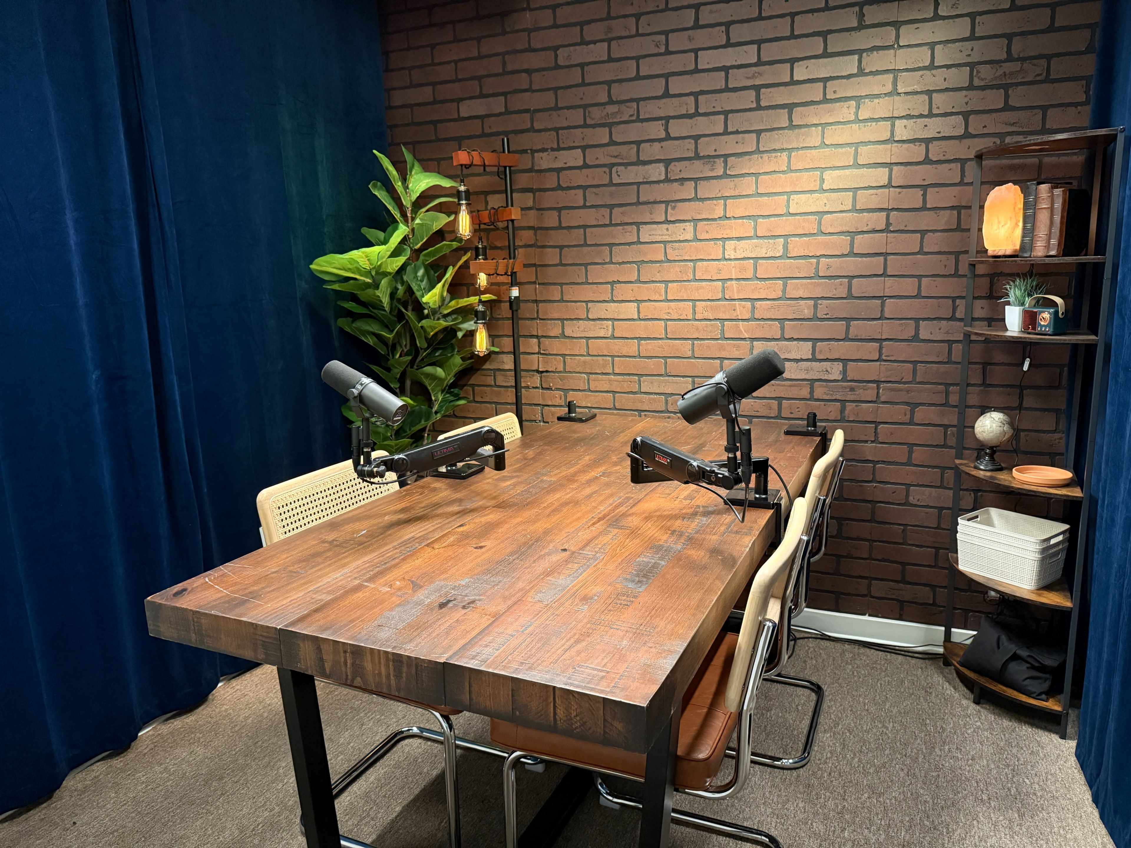 The image shows a podcast recording studio featuring a wooden table with three microphones, surrounded by modern chairs, a brick wall, and green plants.