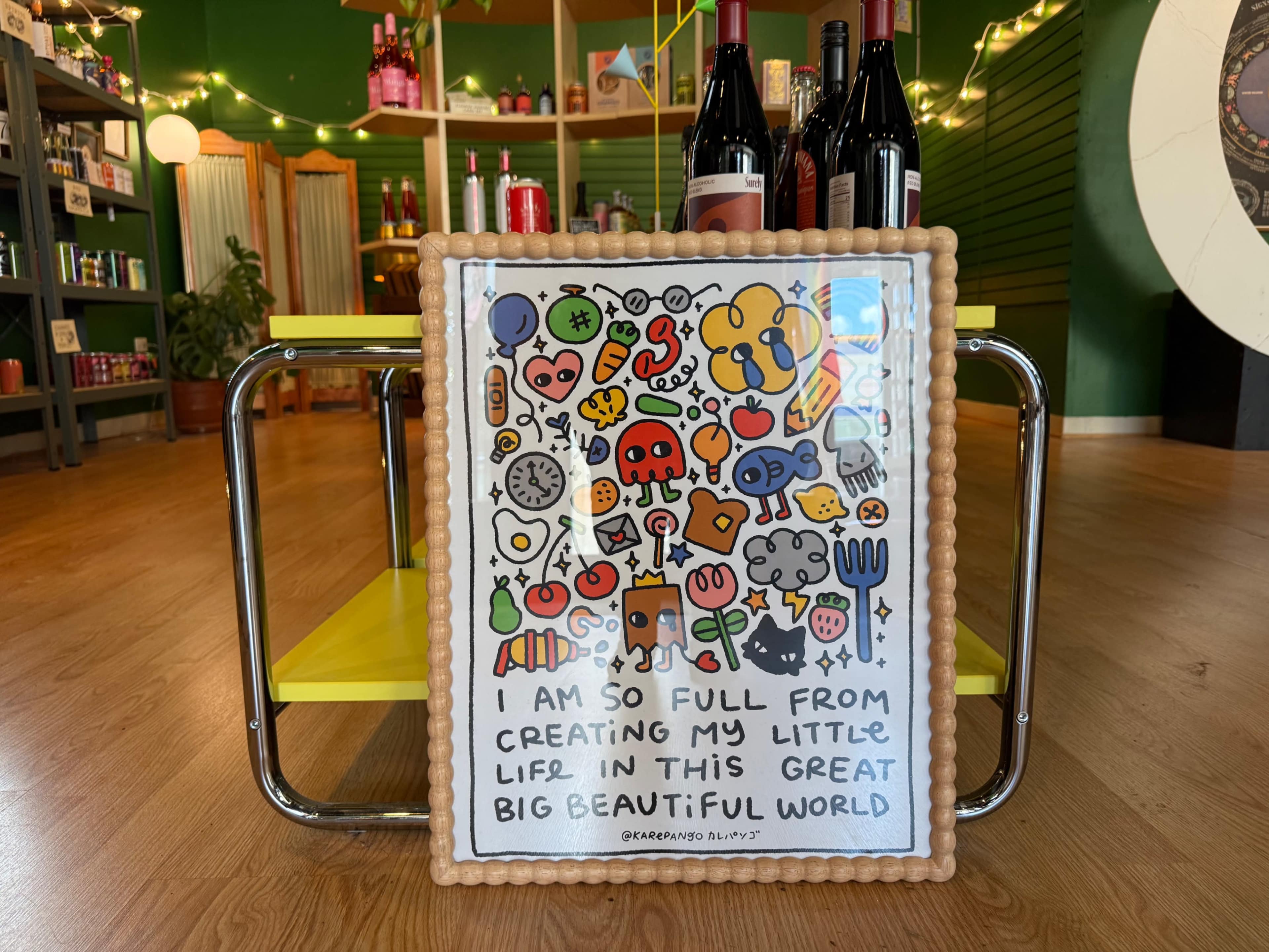 A colorful framed artwork with various illustrations and text sits on a yellow table in a vibrant, well-lit store.