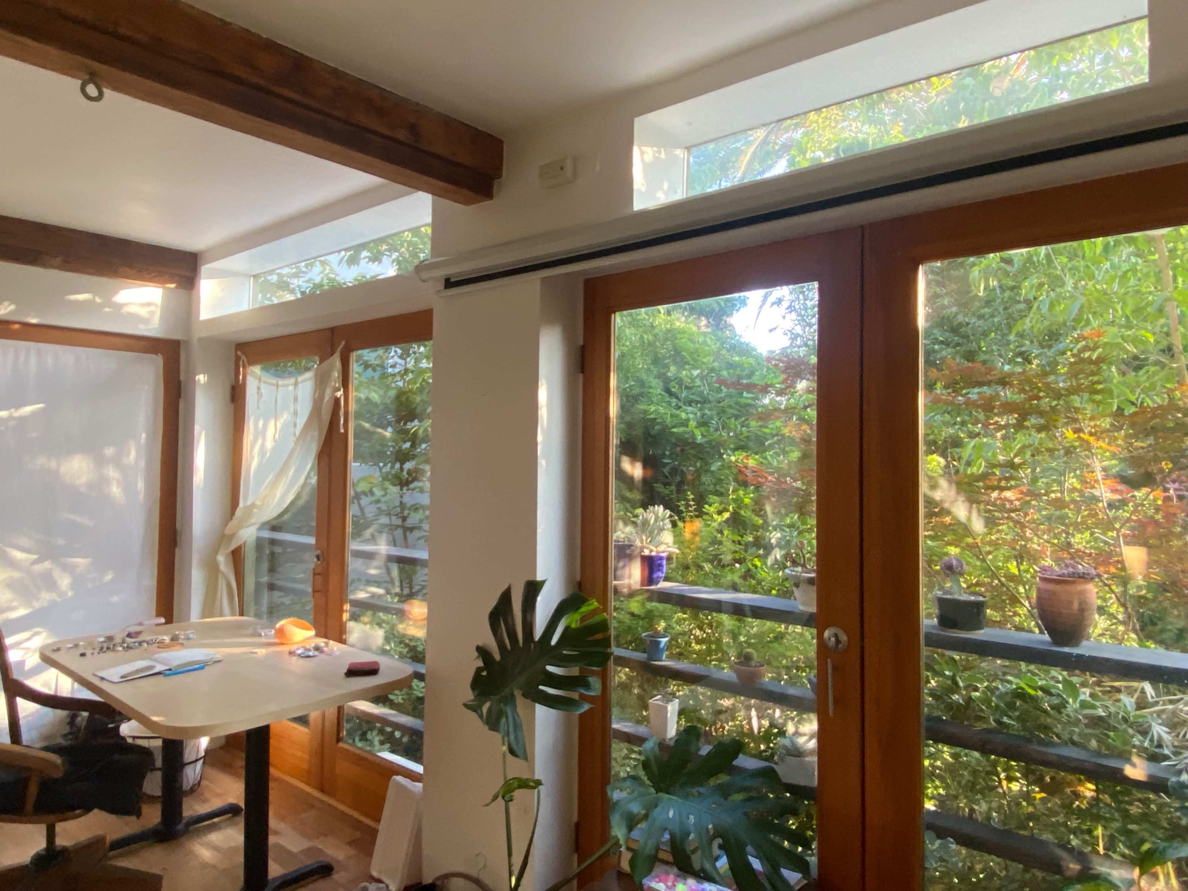 The image shows a bright room with large windows overlooking a garden, featuring a table and chairs inside.