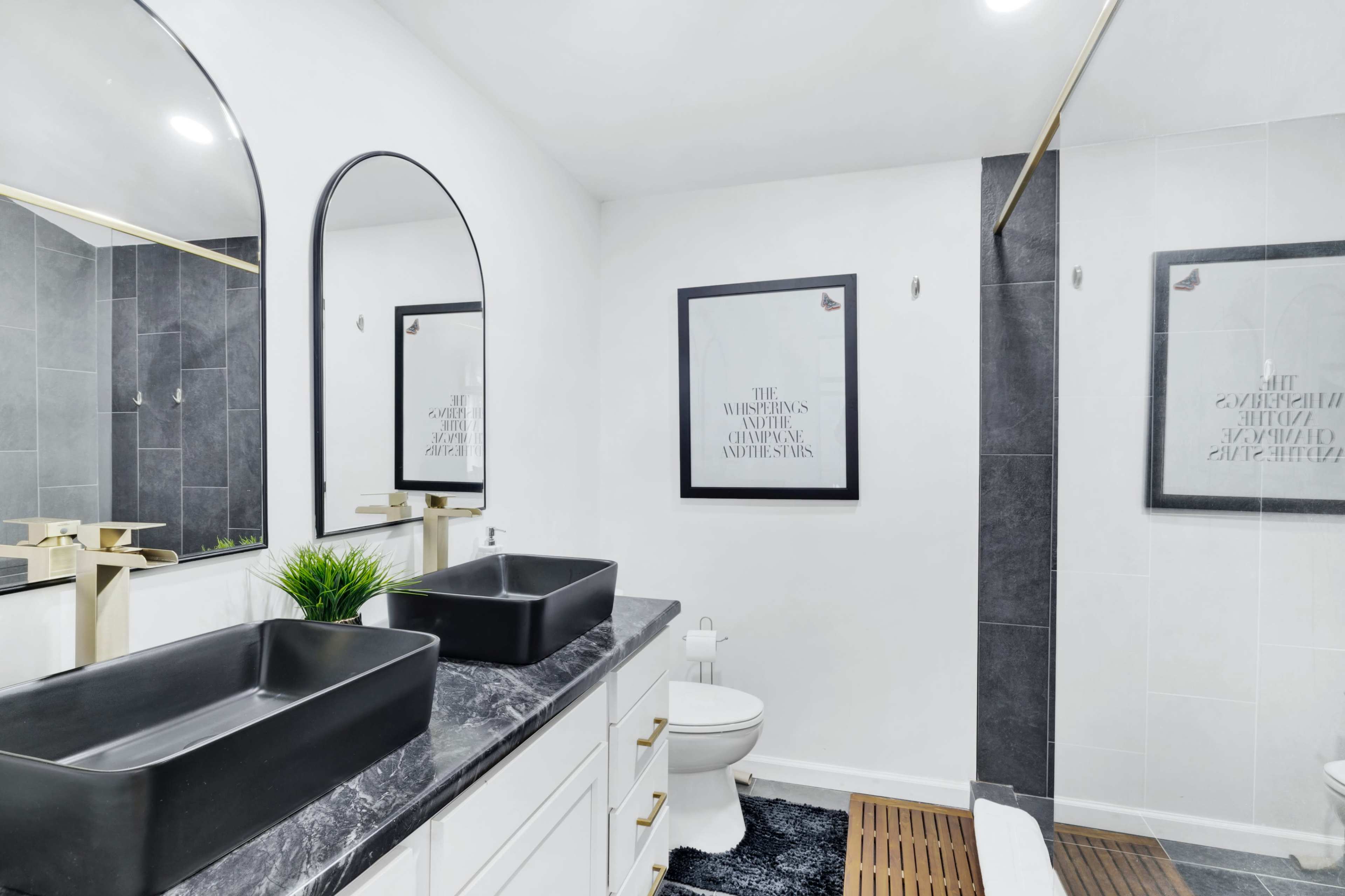 A modern bathroom features two black vessel sinks, a large mirror, a wall-mounted frame, and a tiled shower area.