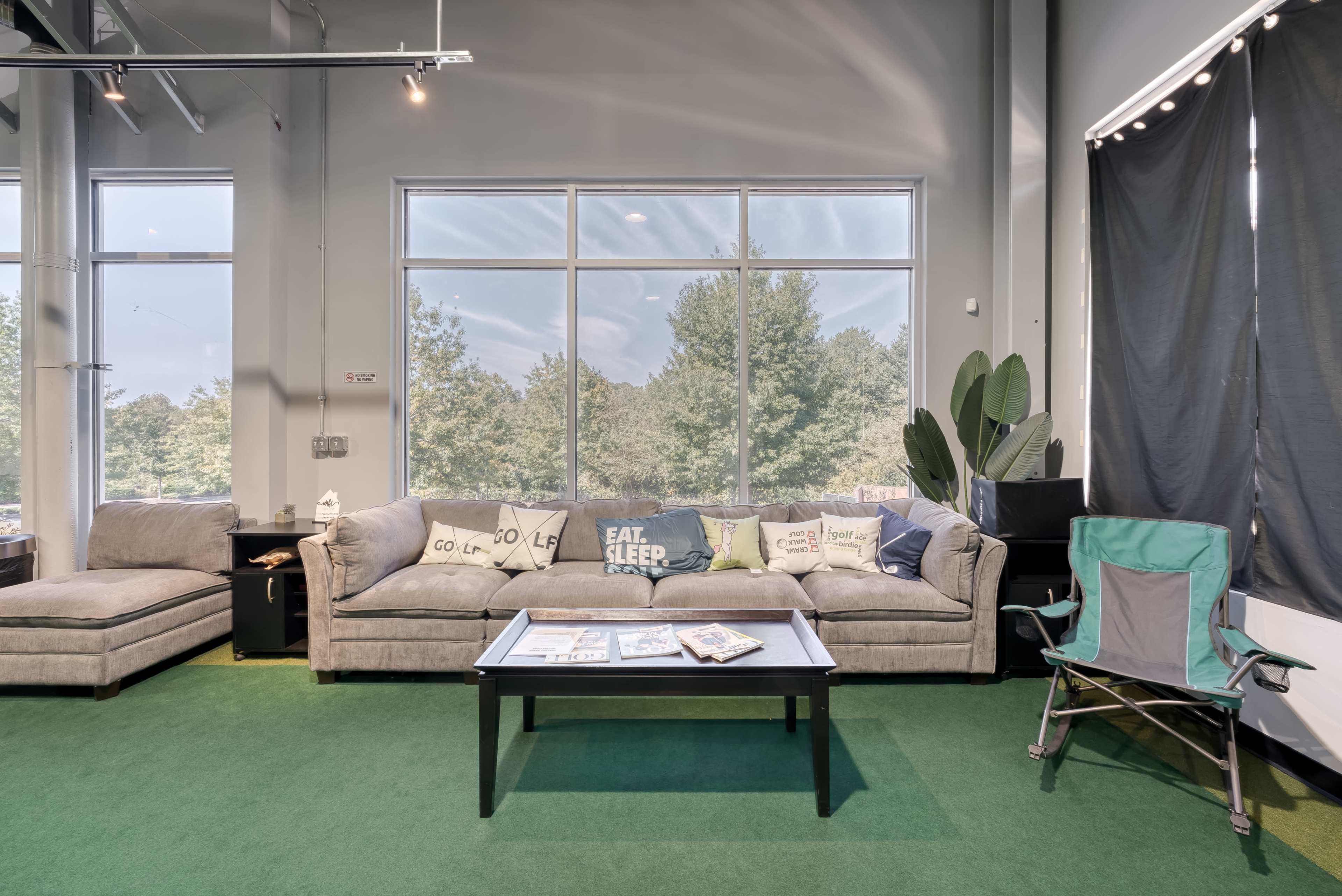 A cozy lounge area featuring a gray sofa with decorative pillows, a black coffee table, a green folding chair, and large windows overlooking trees.