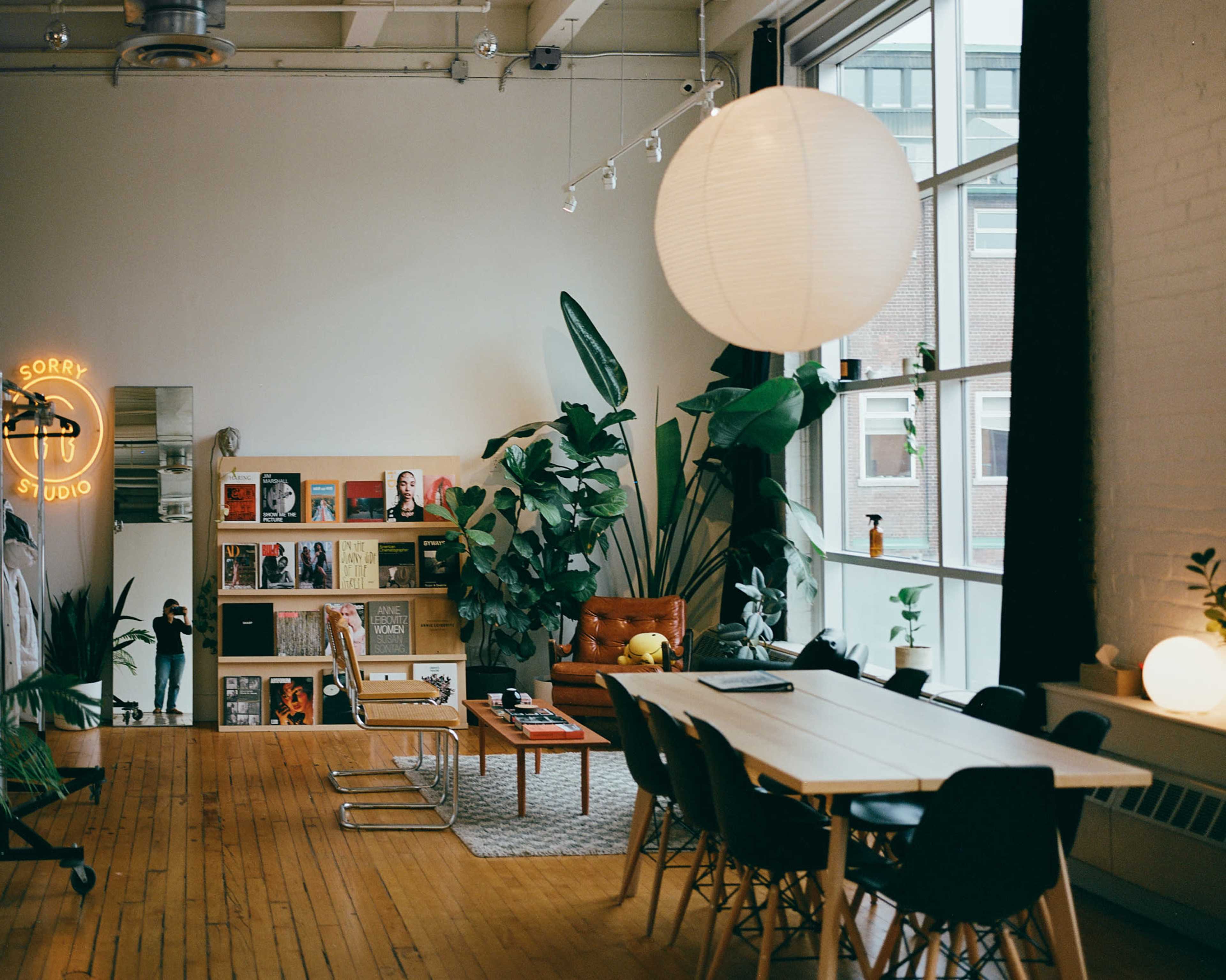 The image shows a modern studio interior featuring a mix of plants, a seating area with a brown leather sofa, a long dining table surrounded by black chairs, and shelves filled with books and decor.