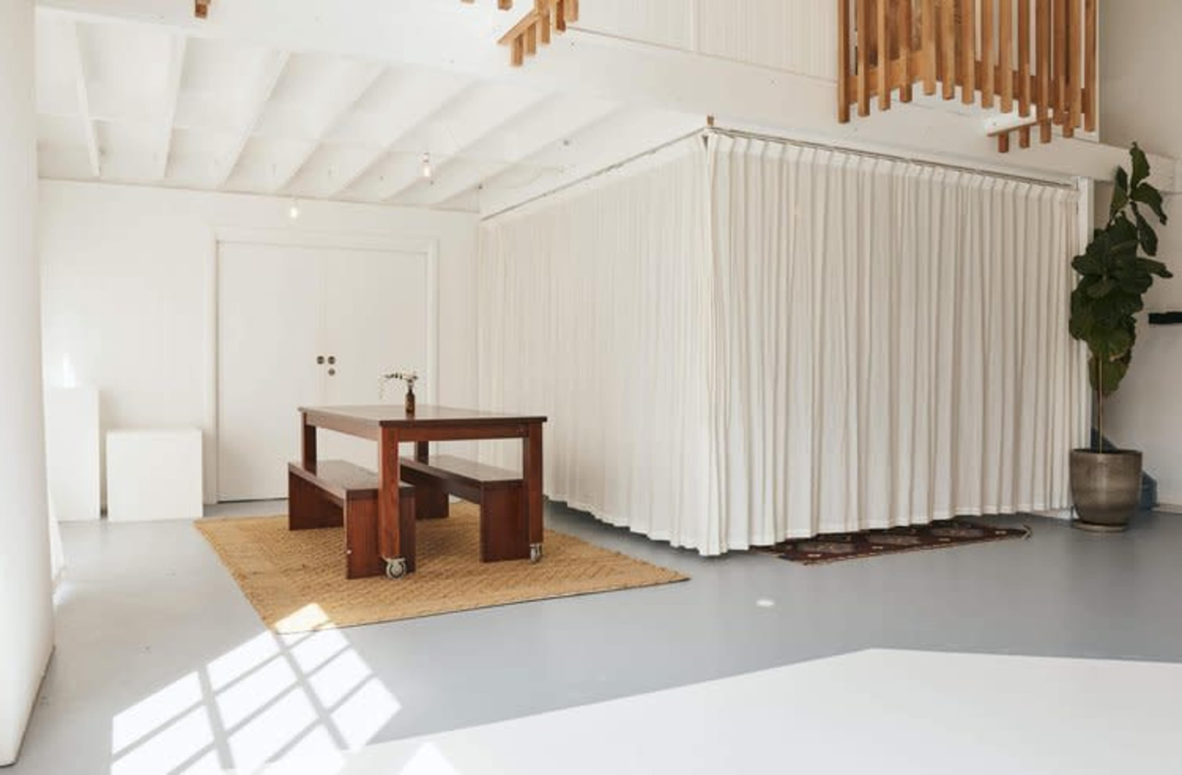 The image shows a bright, minimalist interior space featuring a wooden table on wheels, a woven rug, and a large white curtain separating areas, with a potted plant in the corner.