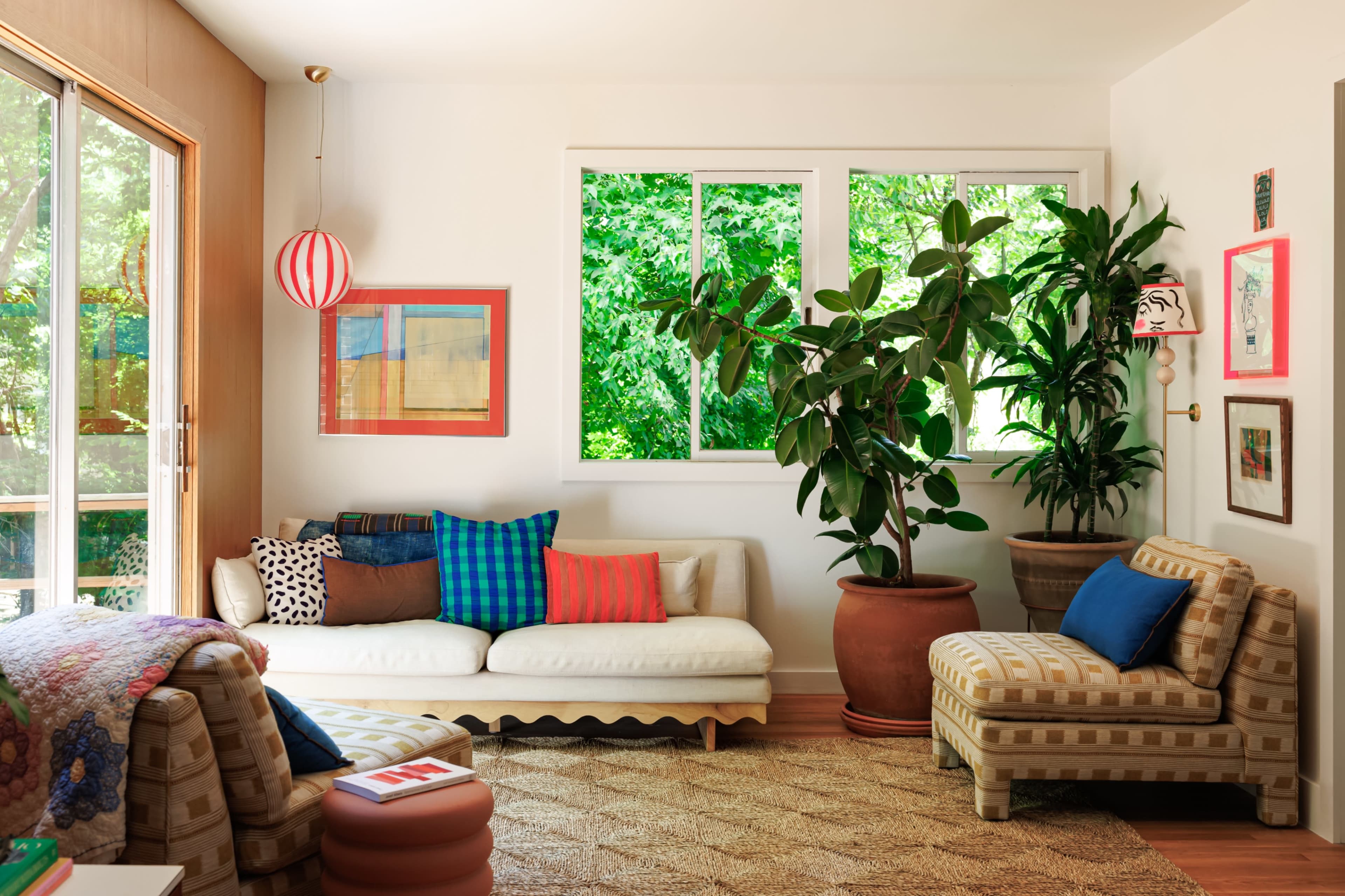The living room features a light-colored sofa and armchair, a large potted plant, and bright wall art beside large windows that overlook greenery.