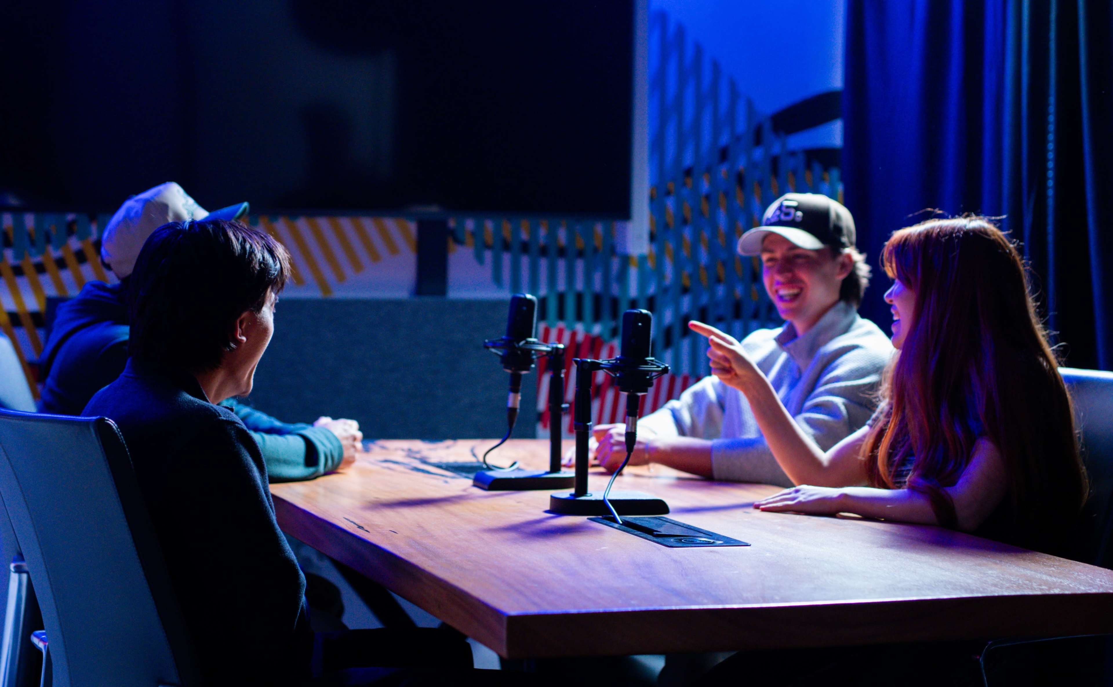 Four people are seated around a wooden table equipped with microphones in a dimly lit recording studio.
