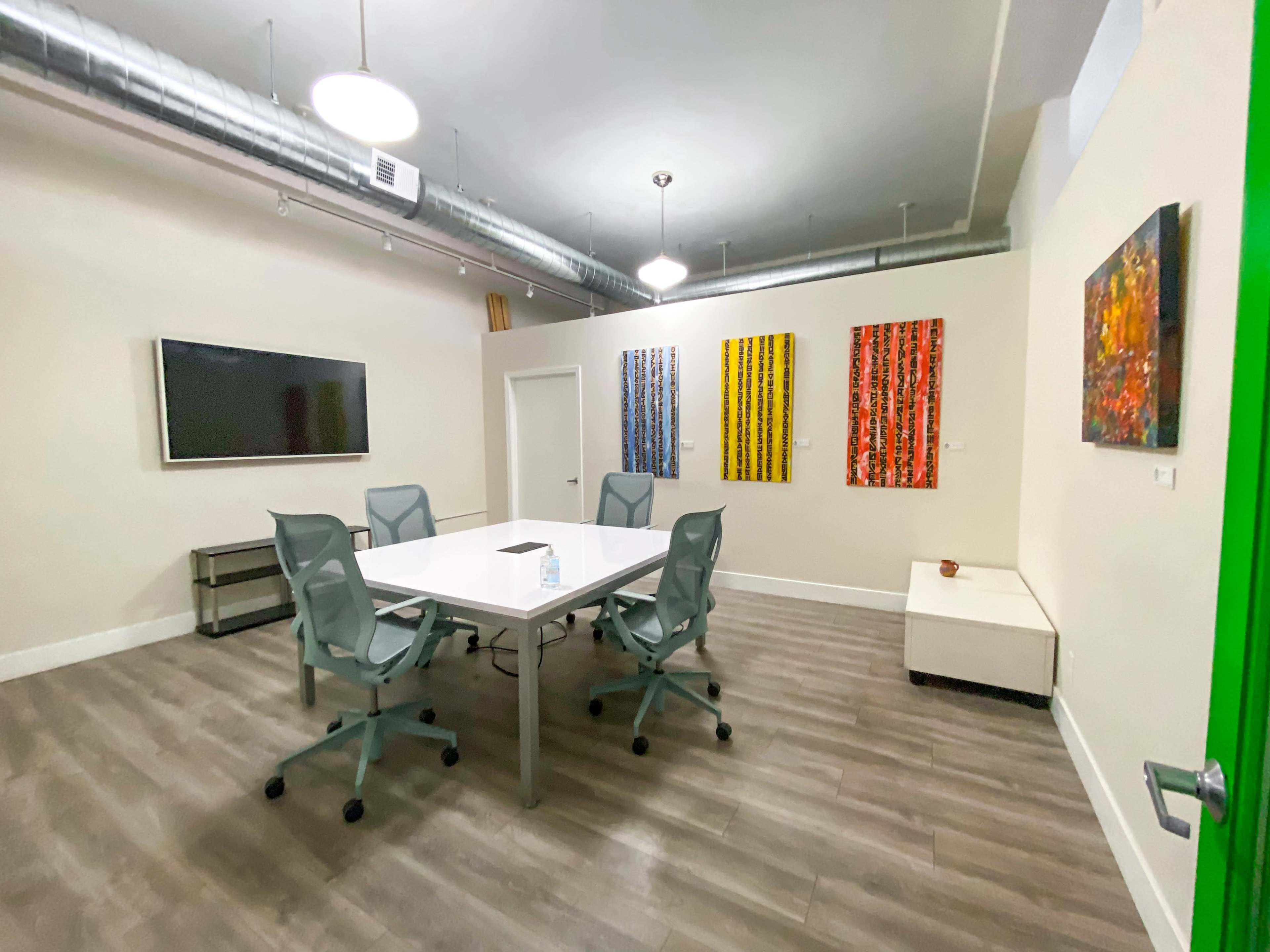 A modern conference room features a large white table surrounded by six ergonomic chairs, with colorful artwork on the walls and a television mounted on one side.